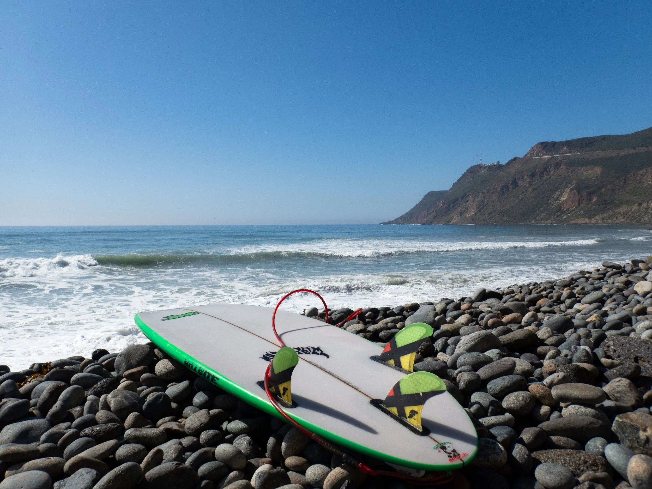 Surfboard with fins resting on a rocky beach, ocean waves and a mountain in the background under a clear blue sky.