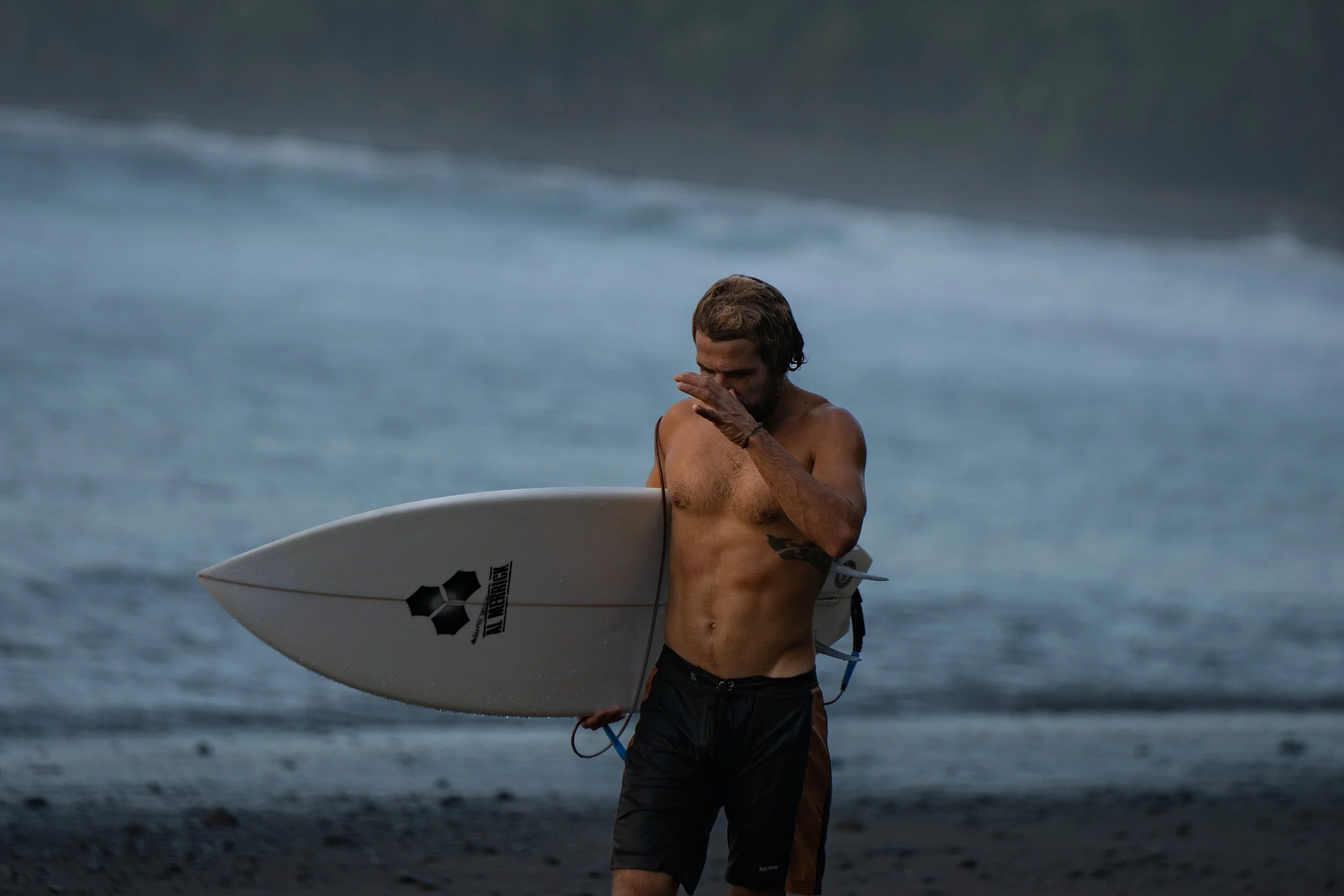 A man with long hair, tan skin, and tattoos on his arm, shirtless, wearing dark shorts, holding a surfboard, walking on the beach with waves in the background.