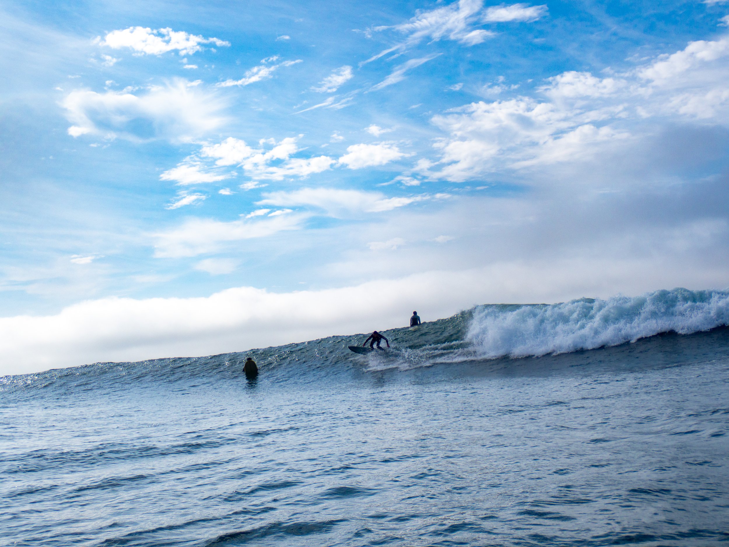 A scene of the ocean with a surfer riding a wave, two others in the water, and a partly cloudy sky.