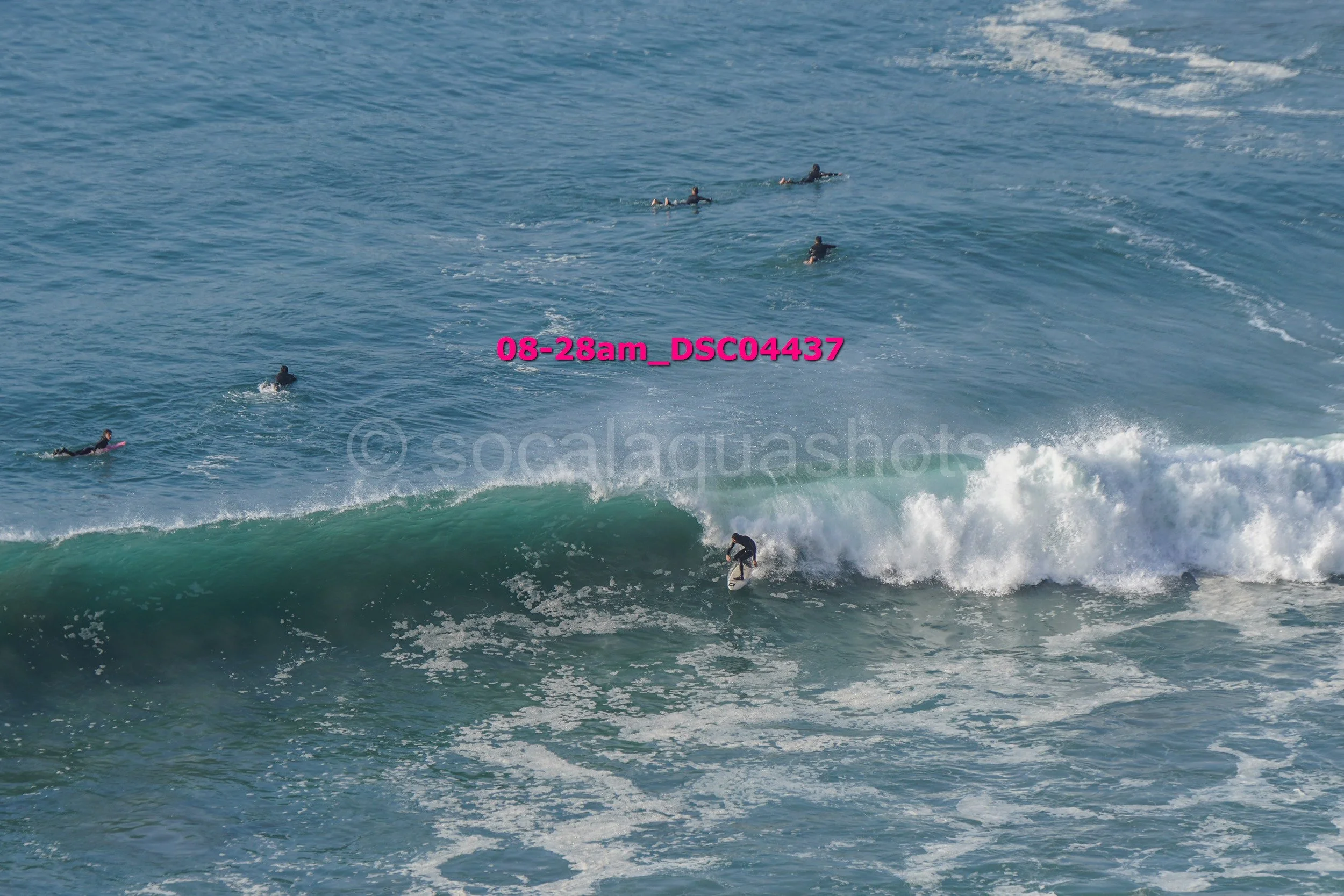 A surfer riding a large wave while several other surfers wait in the water nearby.