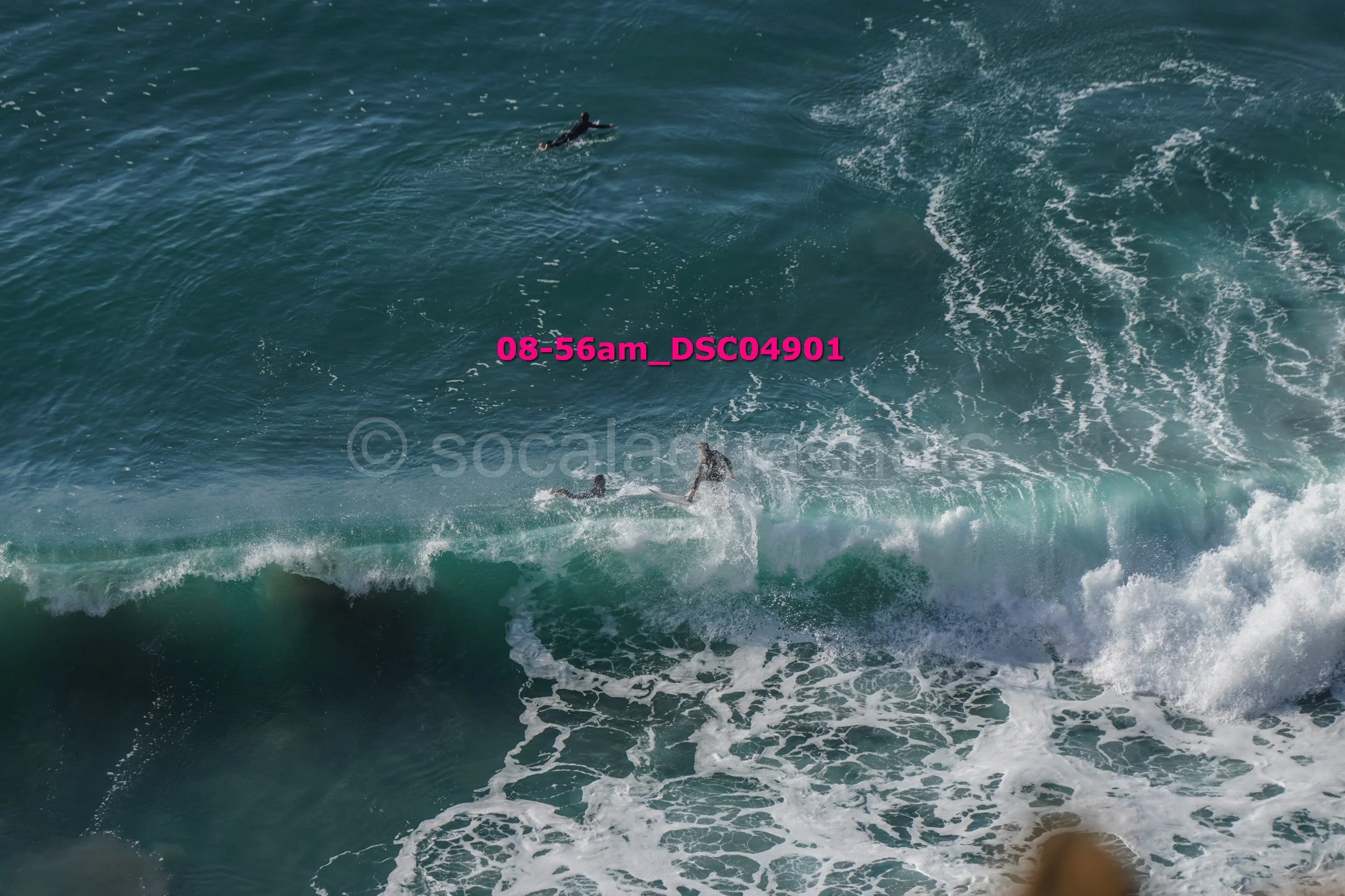 A person surfing on a large ocean wave with two other surfers visible in the distance in the water.