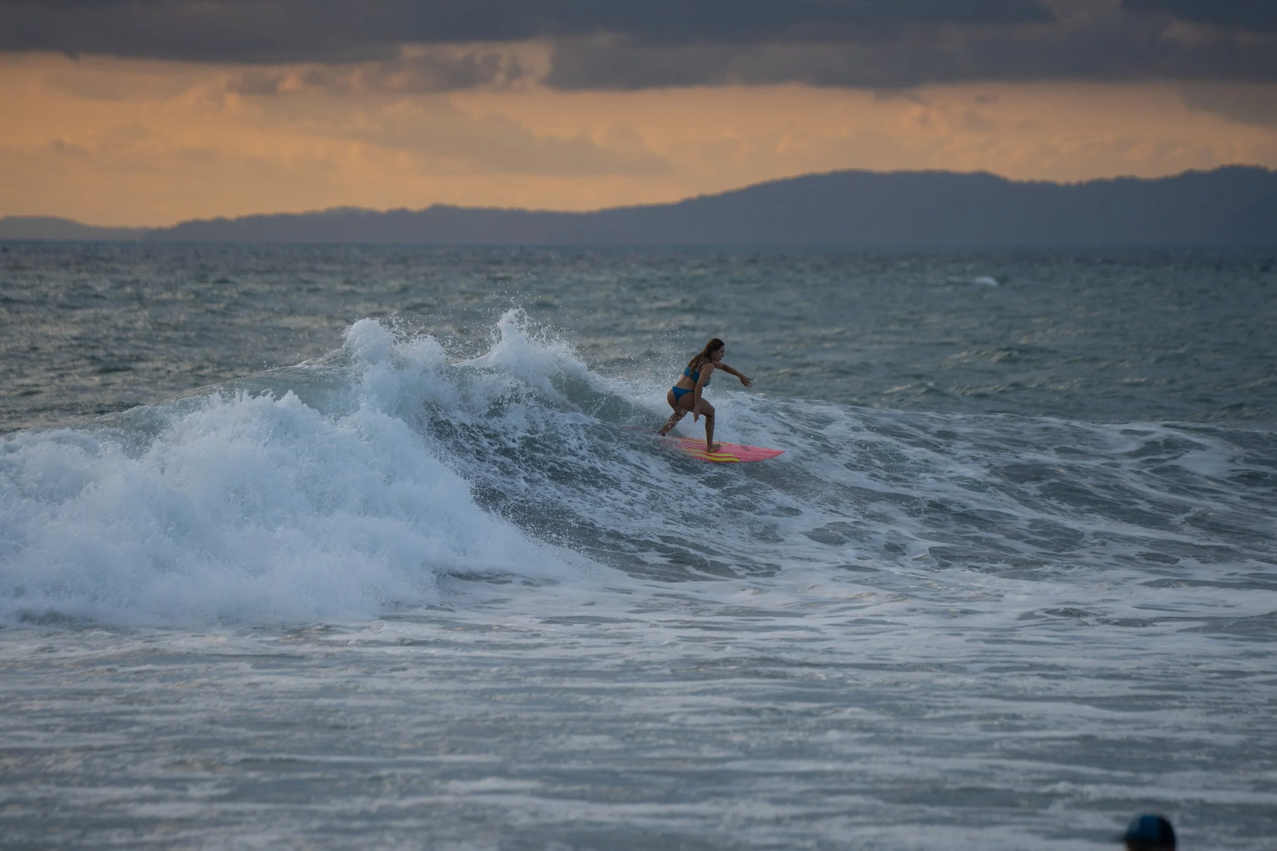 A woman in a blue swimsuit surfing on a pink surfboard on the ocean, with a mountain and cloudy sky in the background.