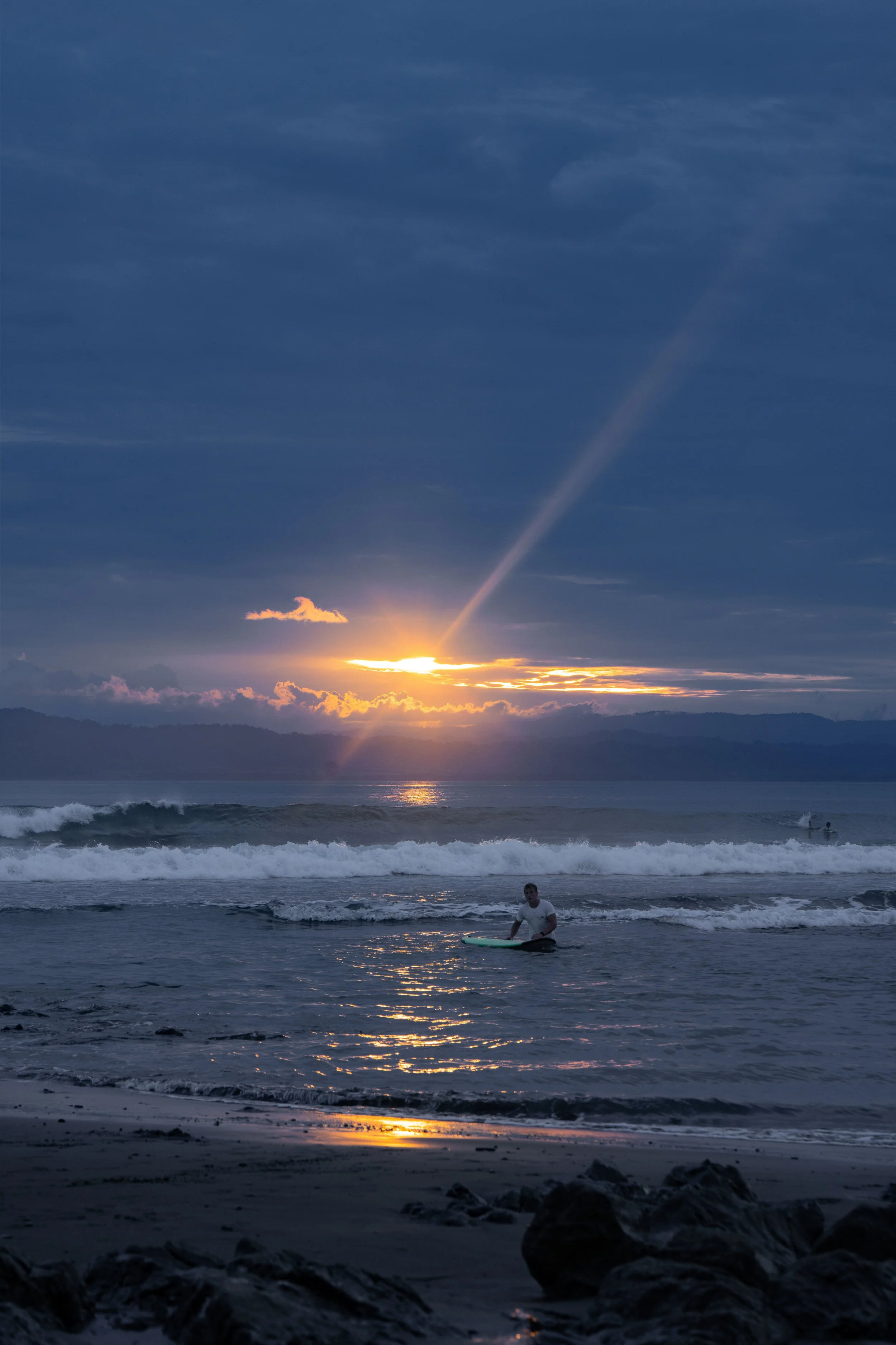 Sunset over the ocean with a visible rainbow and some clouds in the sky. There is a person with a surfboard in the water near the shore, and rocks are visible along the beach.