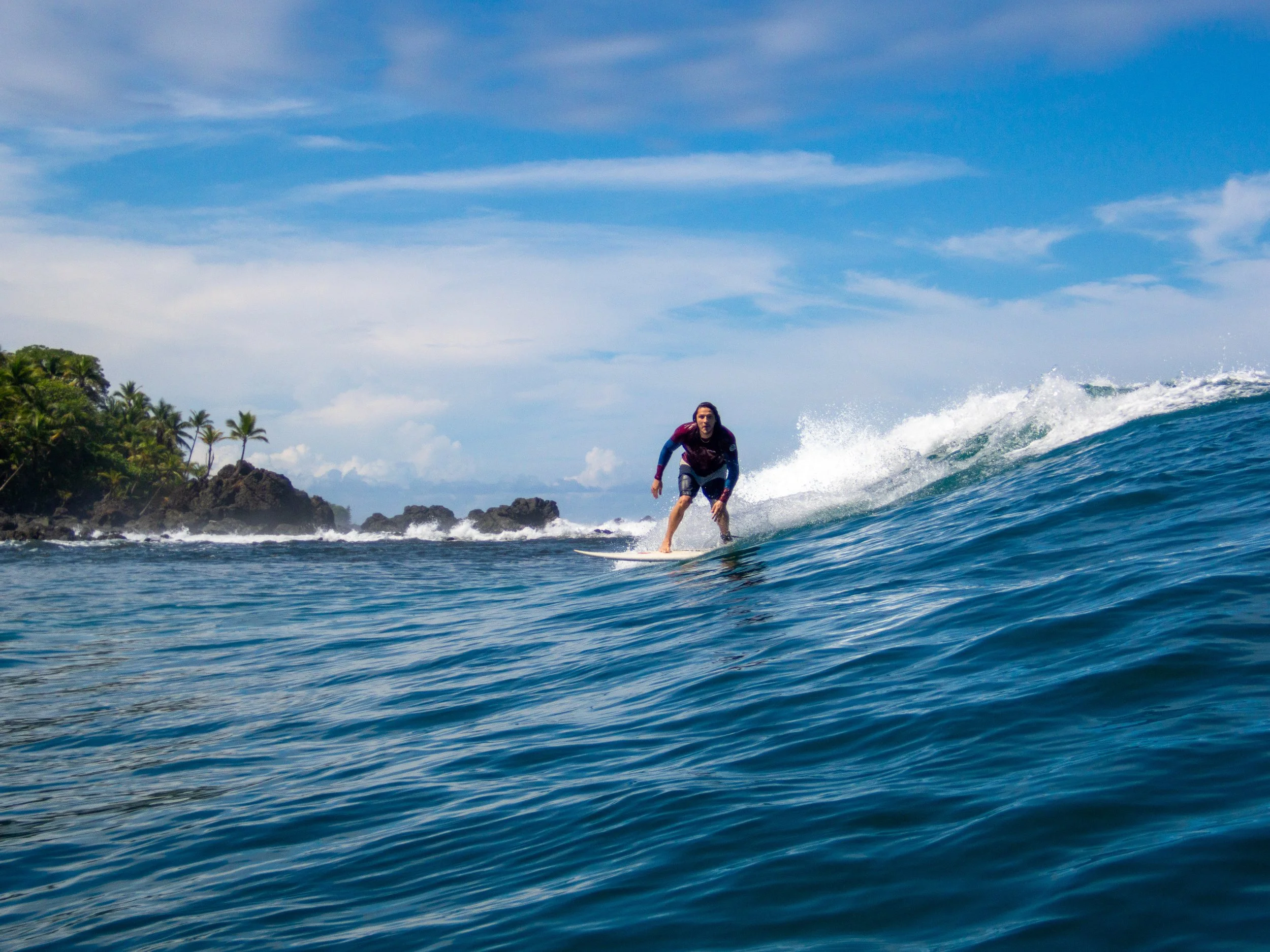 Surfer riding a wave near a tropical coastline with palm trees and rocky cliffs under a blue sky.