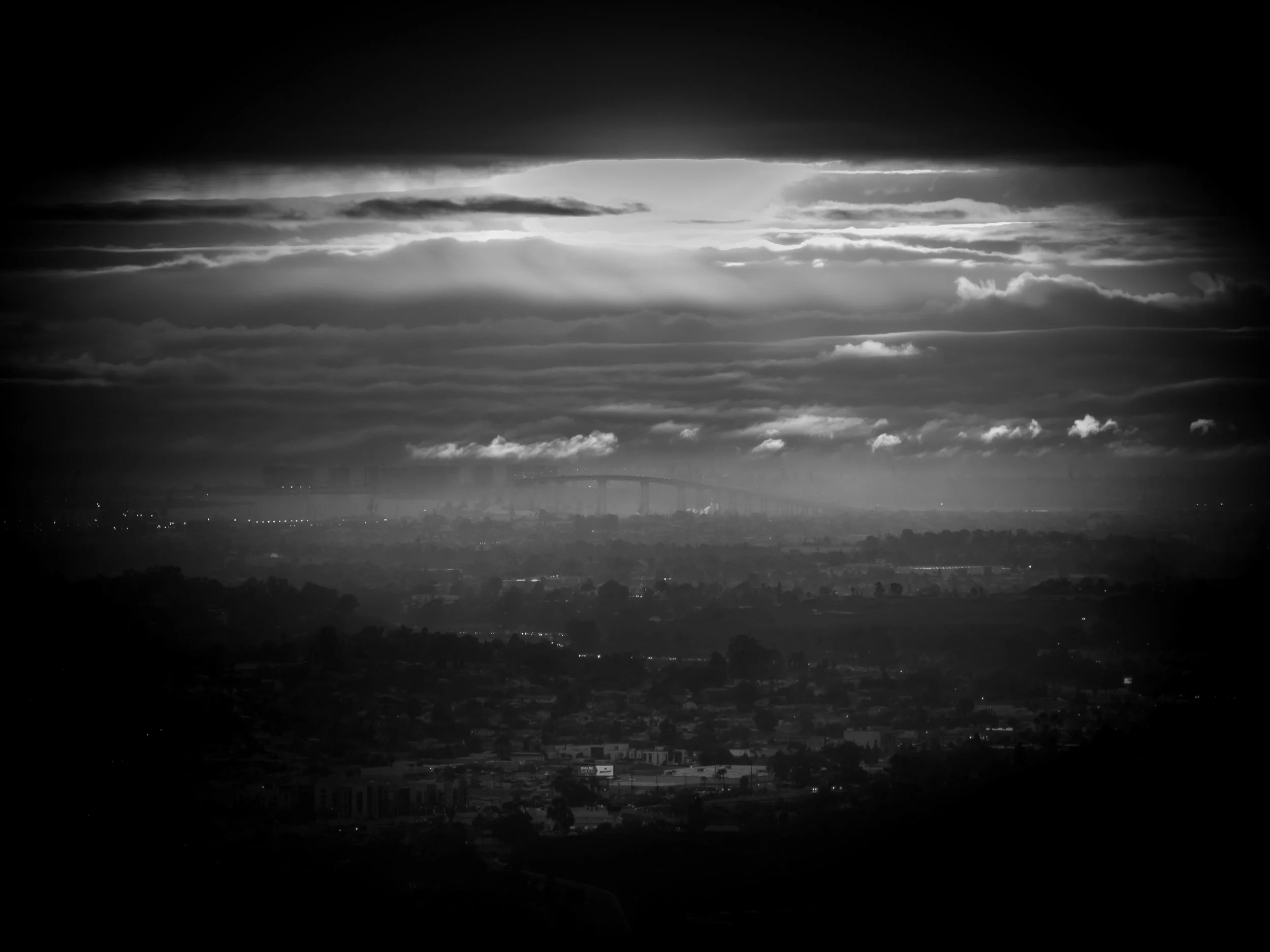 Black and white landscape of a city under cloudy sky with a bridge in the distance