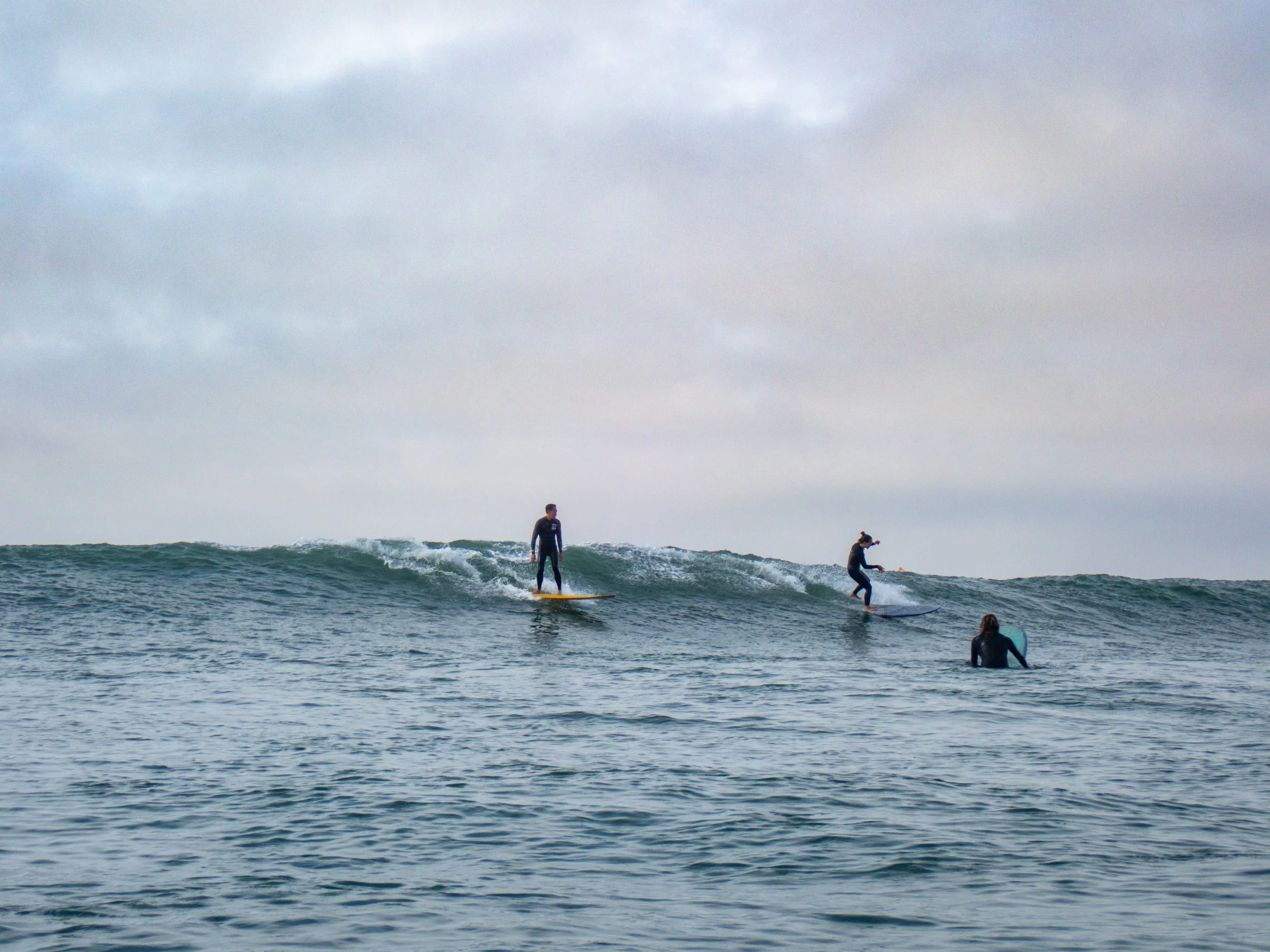 Three surfers on waves in the ocean under a cloudy sky.