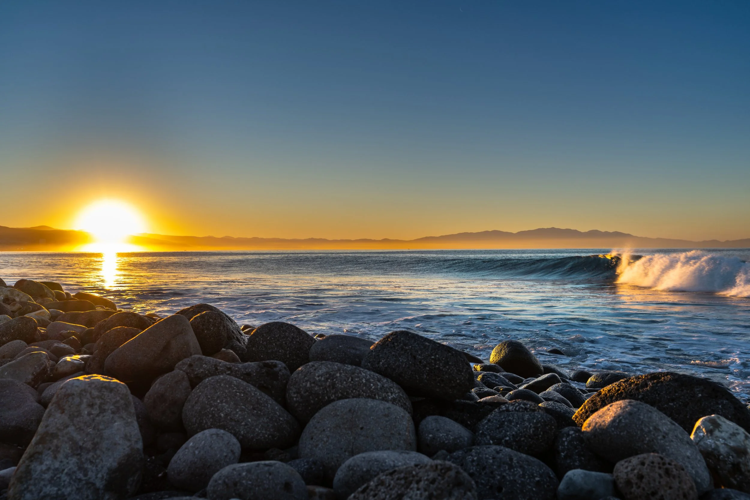 Sunset over the ocean with waves crashing onto a rocky shoreline, mountains in the distance