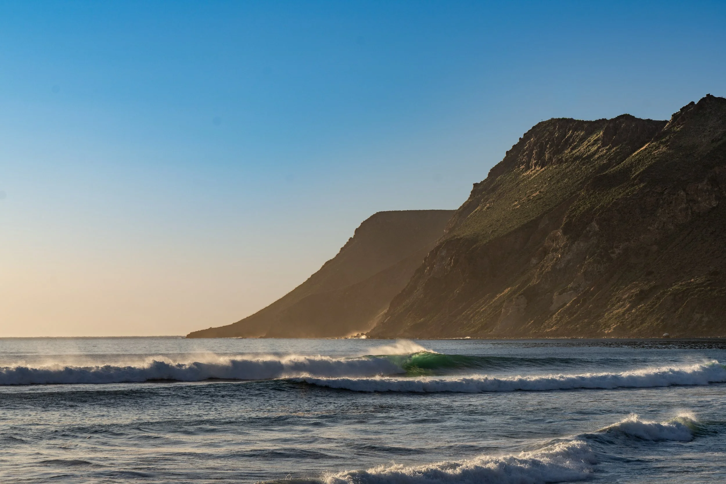 Sunset over ocean with waves and green cliffs in the background.