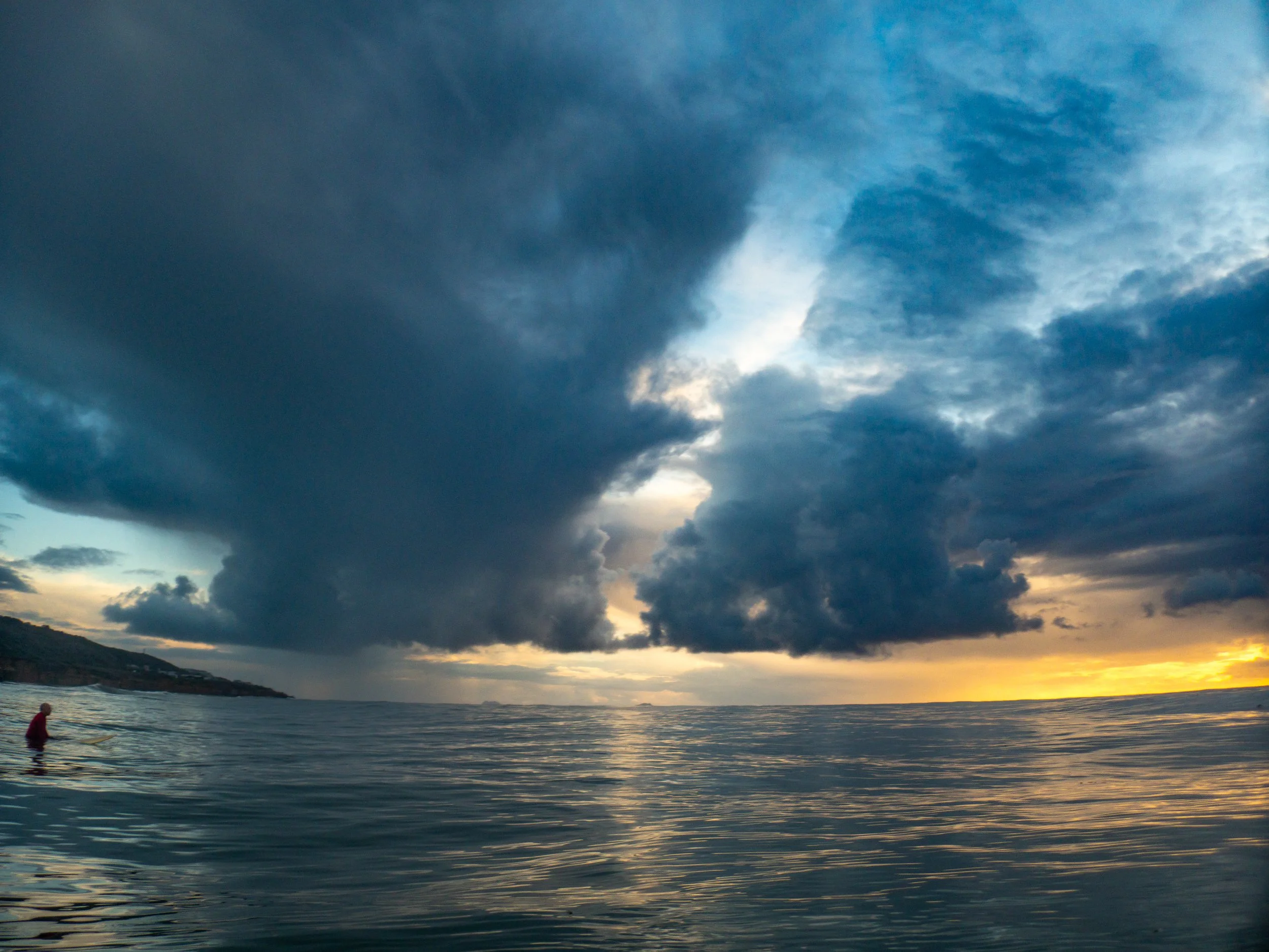 A person in a wetsuit standing in the ocean water near the shoreline under a dramatic cloudy sky during sunset.