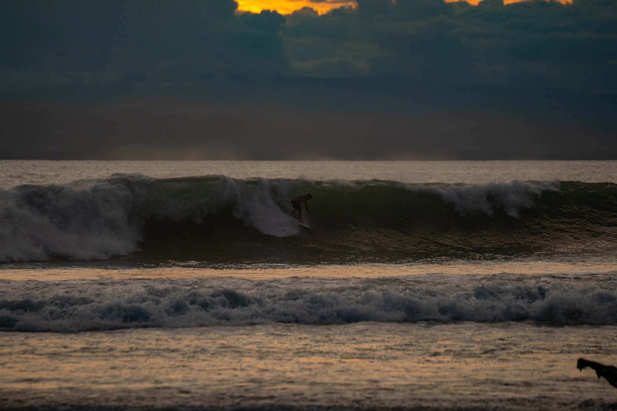A person surfing on a wave during sunset with dark clouds in the sky.