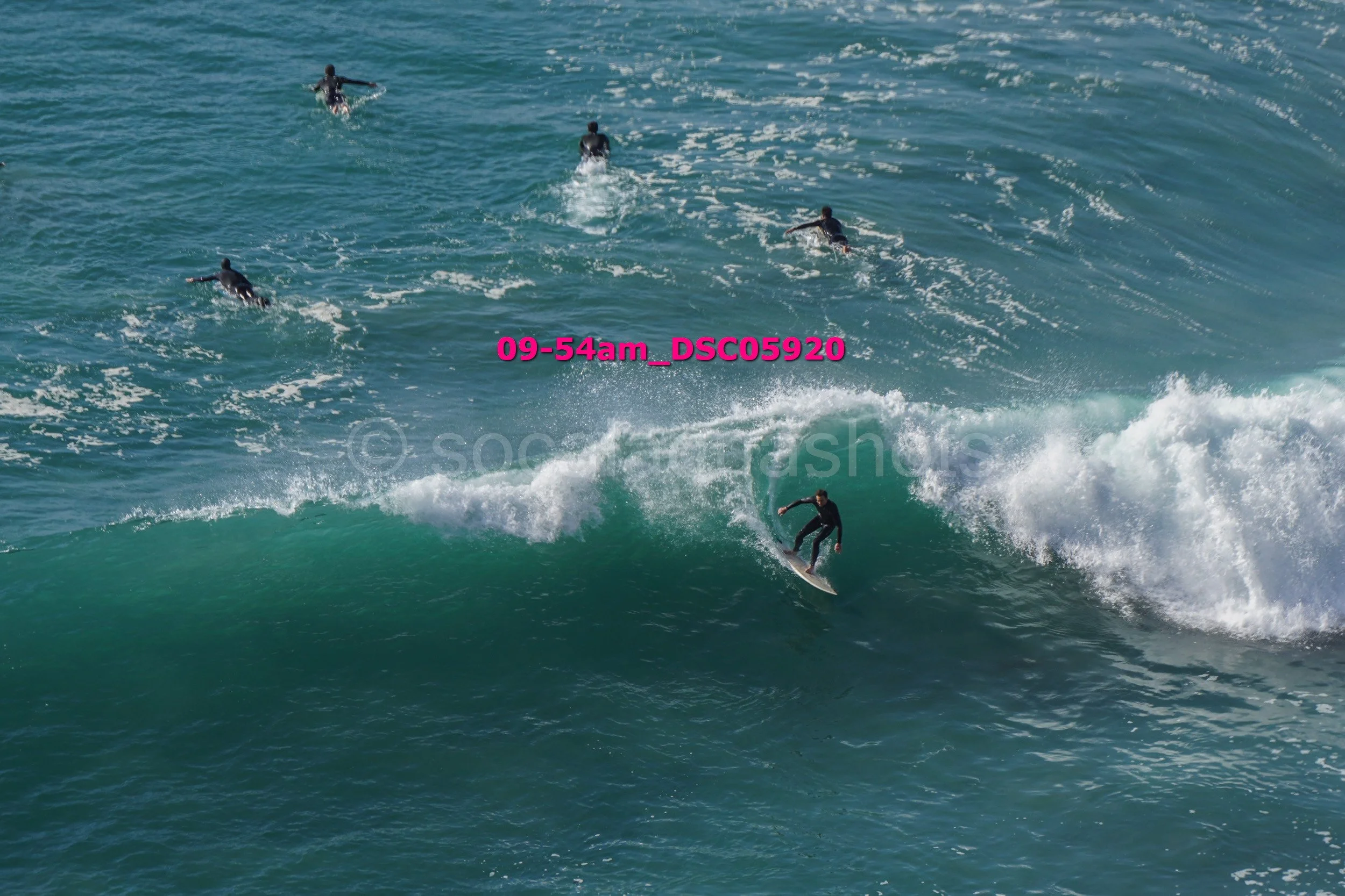 Surfer riding a wave with several people swimming or surfing in the background in the ocean.