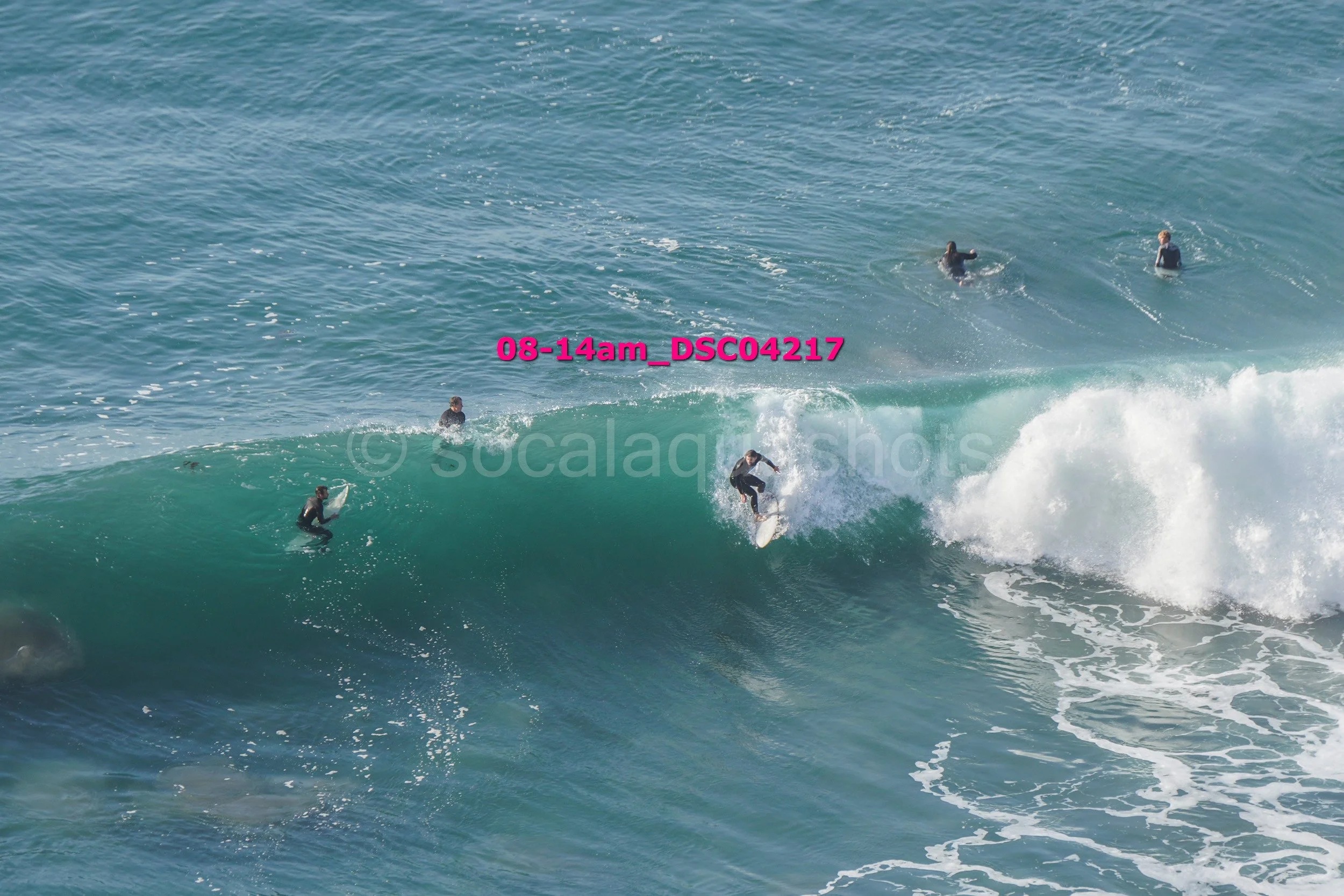 Surfers riding a large wave in the ocean with several other surfers in the water nearby.