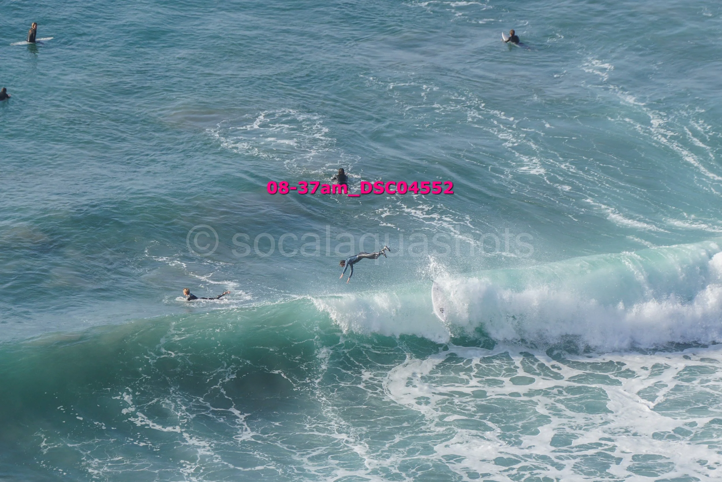 Surfer falling off the surfboard in the waves at the beach, with several other surfers in the water.