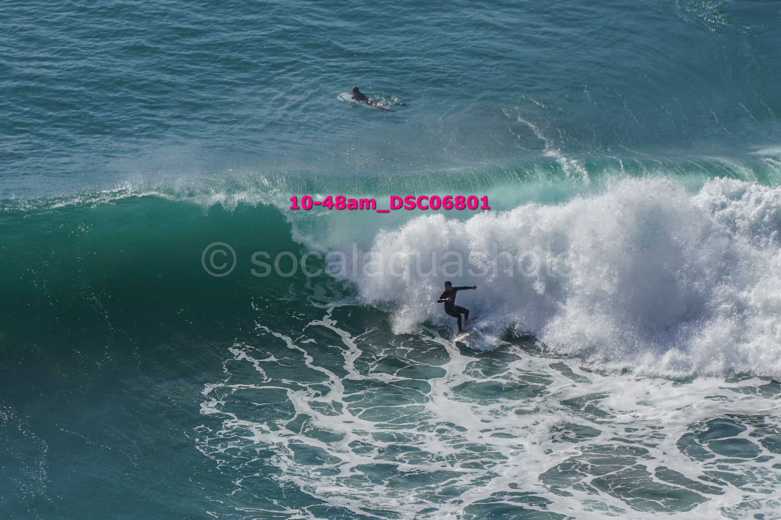 A person surfing on a large wave in the ocean with another surfer swimming in the background.