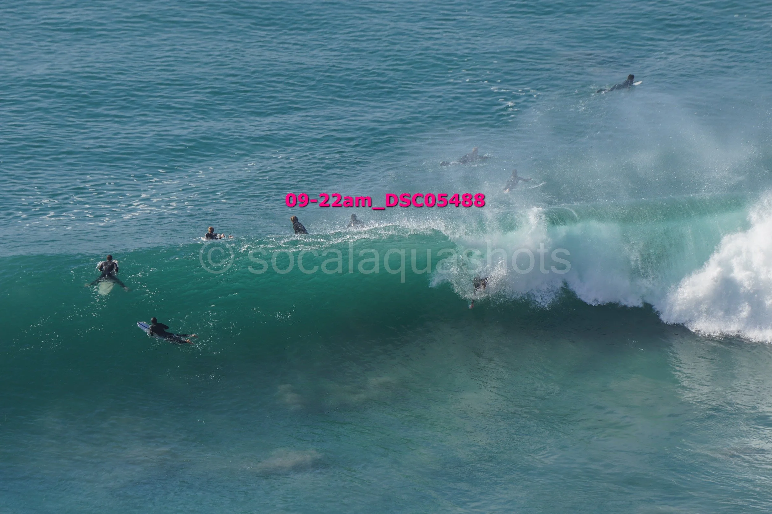 Surfers catching a wave in the ocean, with some surfers riding the wave and others waiting in the water, under clear skies.