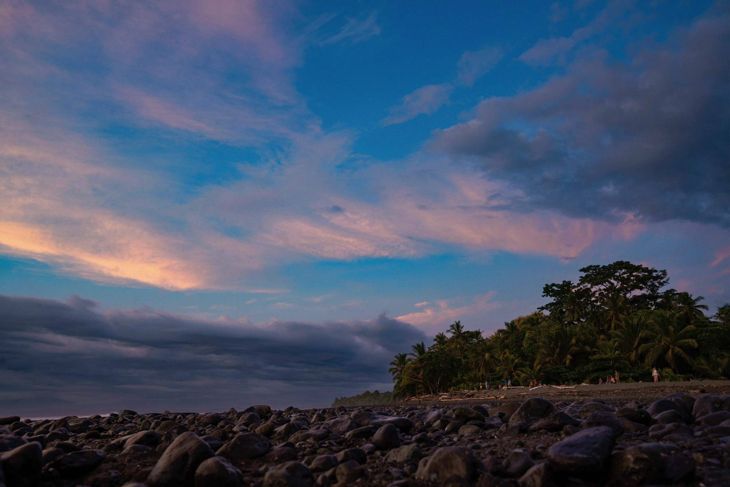 A sunset or evening view of a rocky beach with a forested area and people gathered near the shore, under a partly cloudy sky with pink and blue hues.