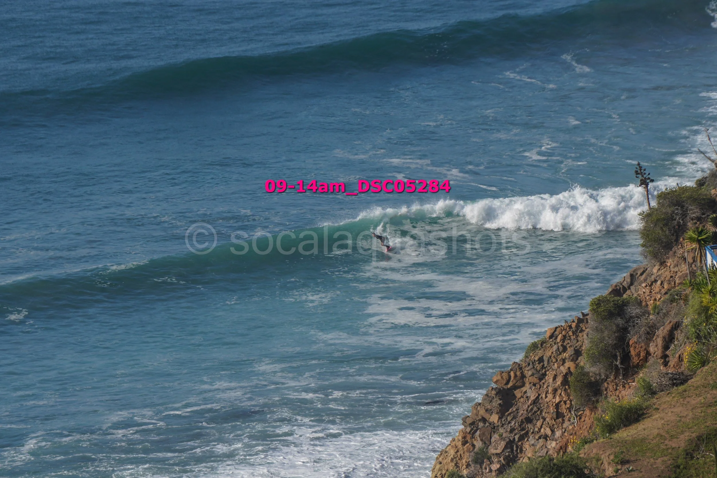 A person surfing on a wave in the ocean near the rocky shoreline with greenery and a flag on a pole.