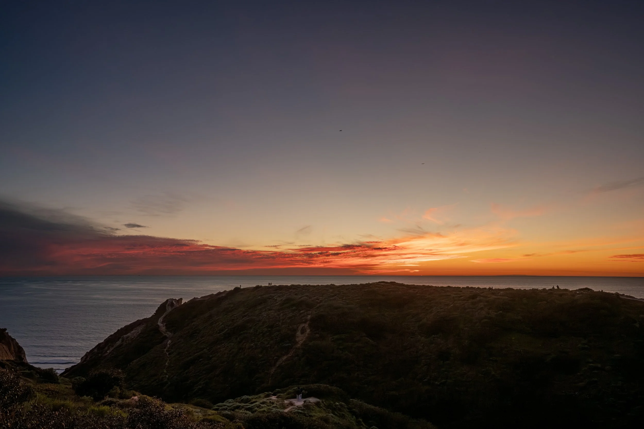 Scenic view of a sunset over the ocean with a hilly coastline in the foreground and a sky filled with orange, pink, and purple hues.