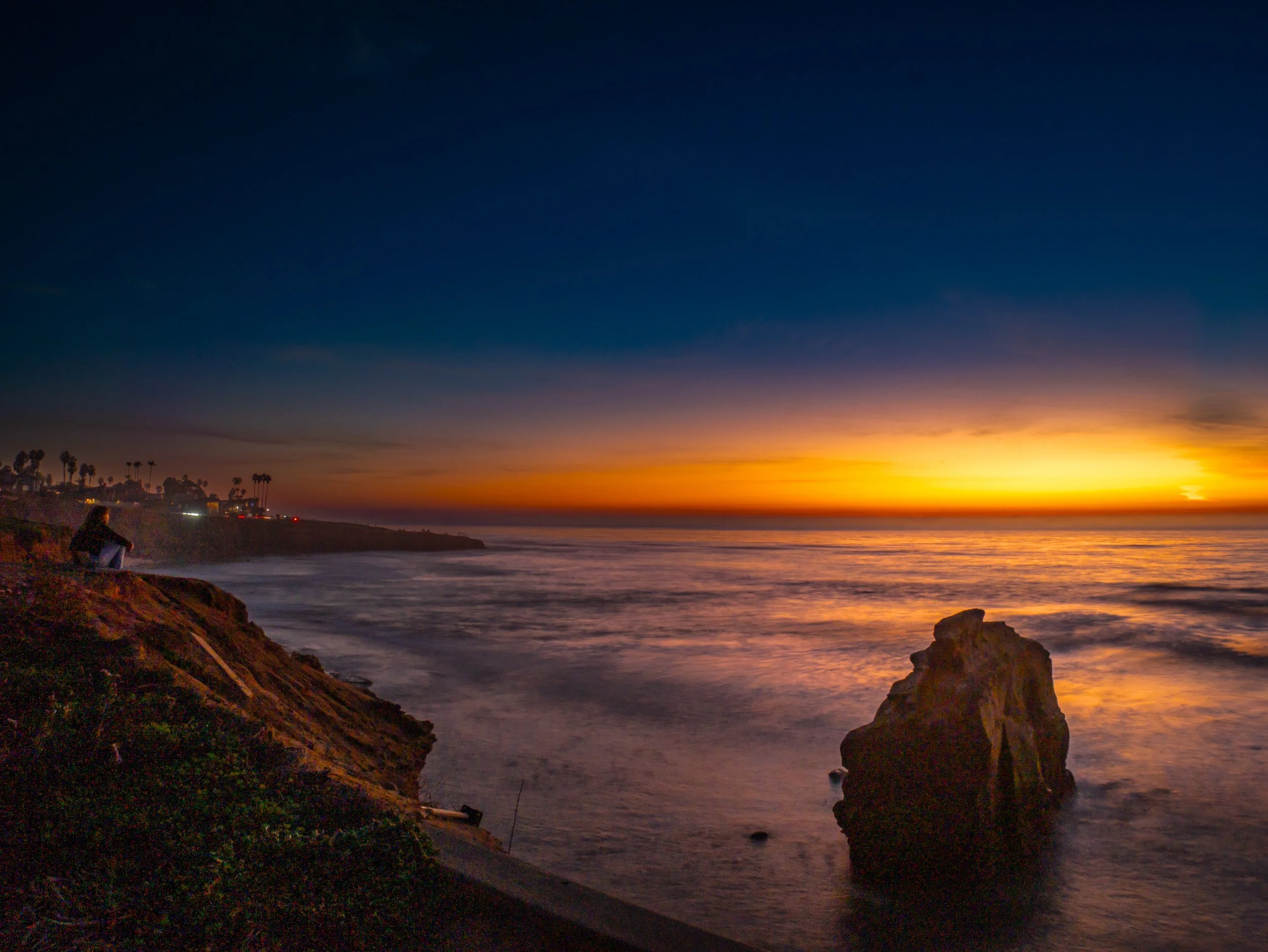 A person sitting on a rocky cliff overlooking the ocean during sunset, with colorful sky and distant shoreline.