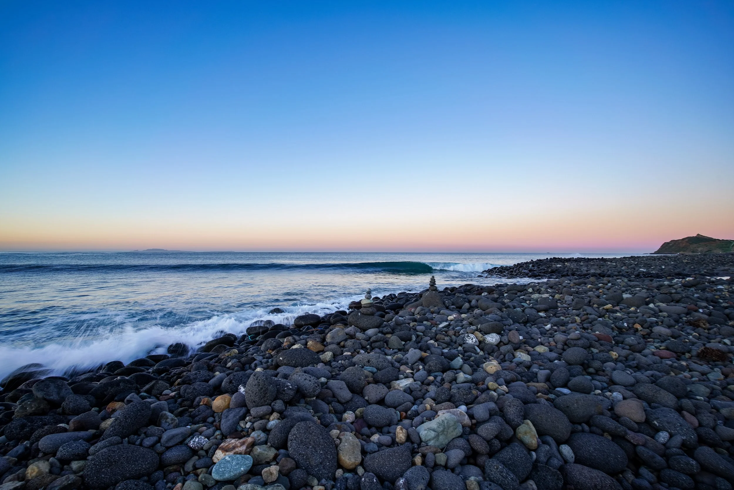A rocky beach at sunset with a few stone cairns, gentle waves, and a distant landmass on the horizon.
