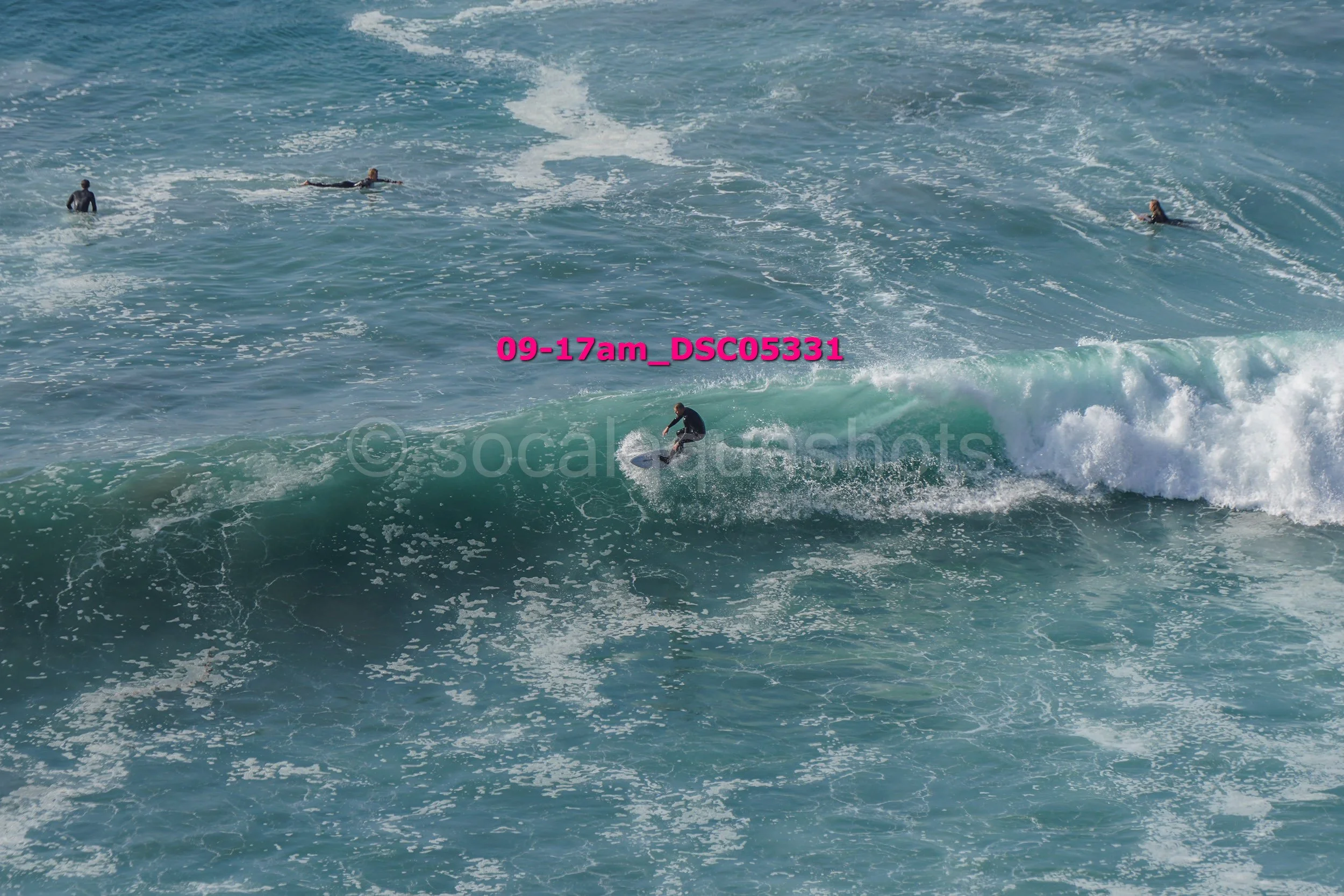 A person in a wetsuit surfing on a large wave with three other surfers in the water nearby.