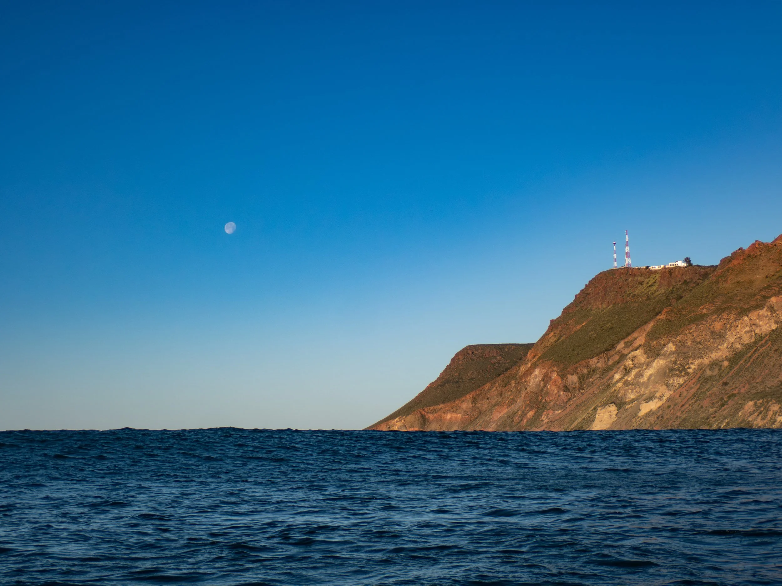 View of a clear blue sky over the ocean with a large hill or cliff on the right side, topped with communication towers and some buildings, with the moon visible in the sky.