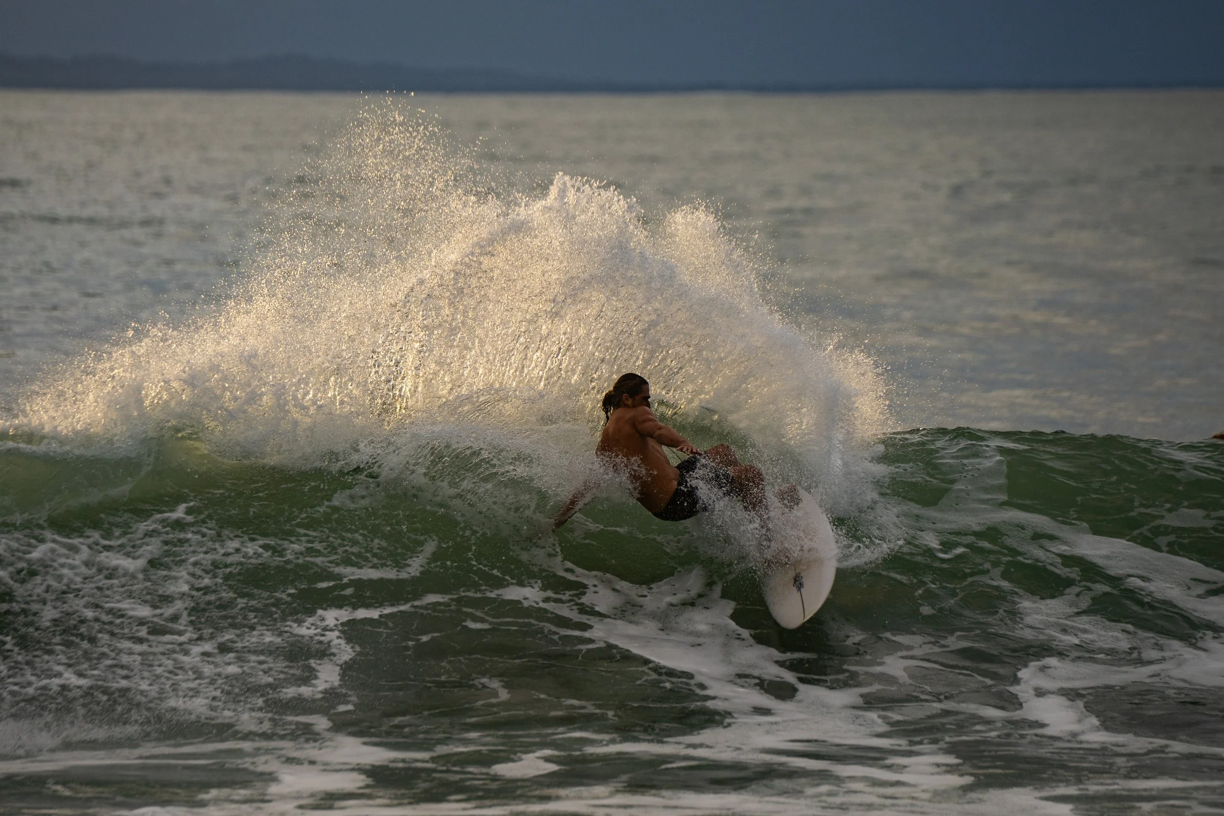 A man windsurfing on the ocean at sunset, creating a spray of water as he rides a wave.