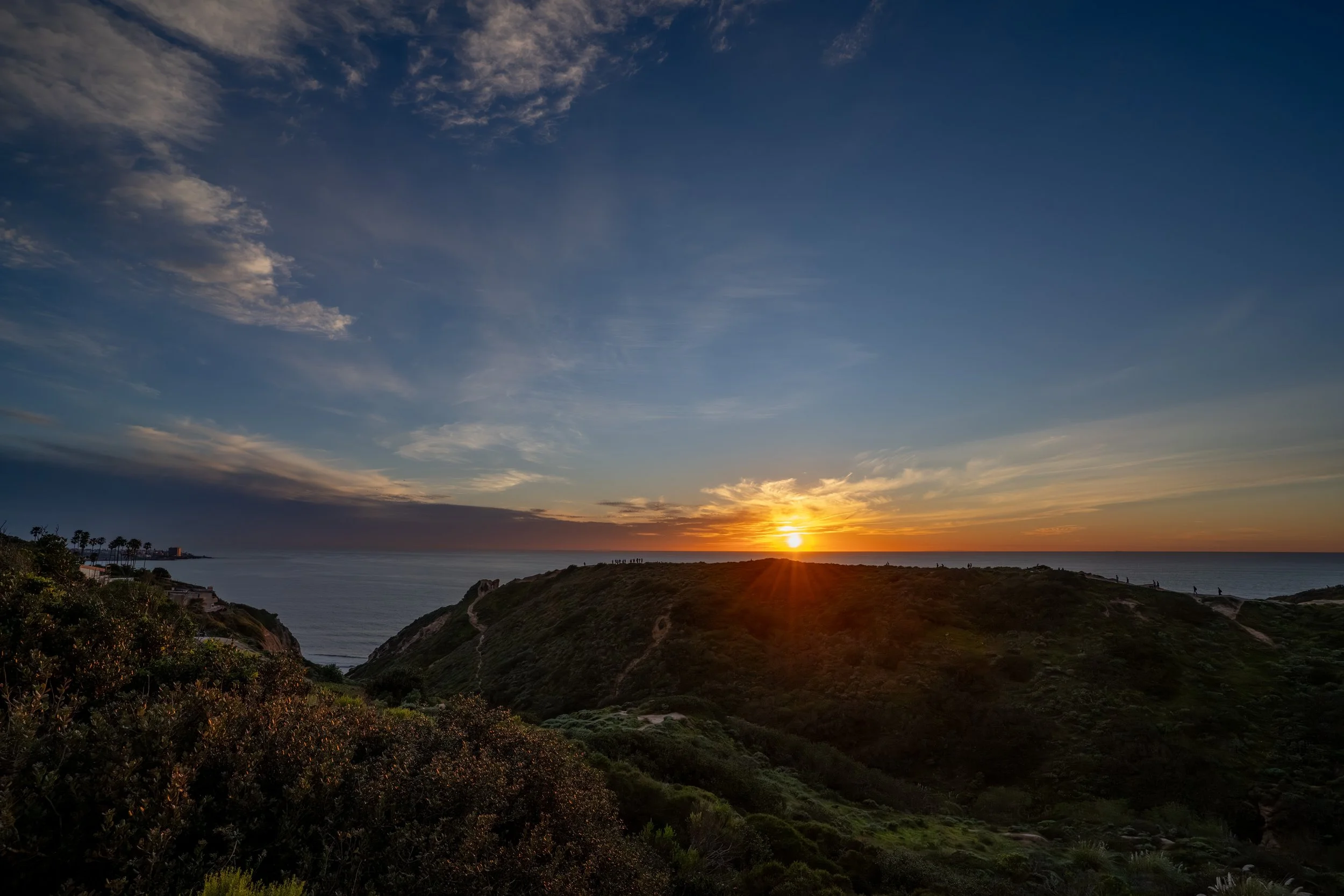 Sunset over a coastal landscape with rolling hills covered in vegetation, a few people silhouetted on the horizon, and a partly cloudy sky with shades of blue, orange, and yellow.