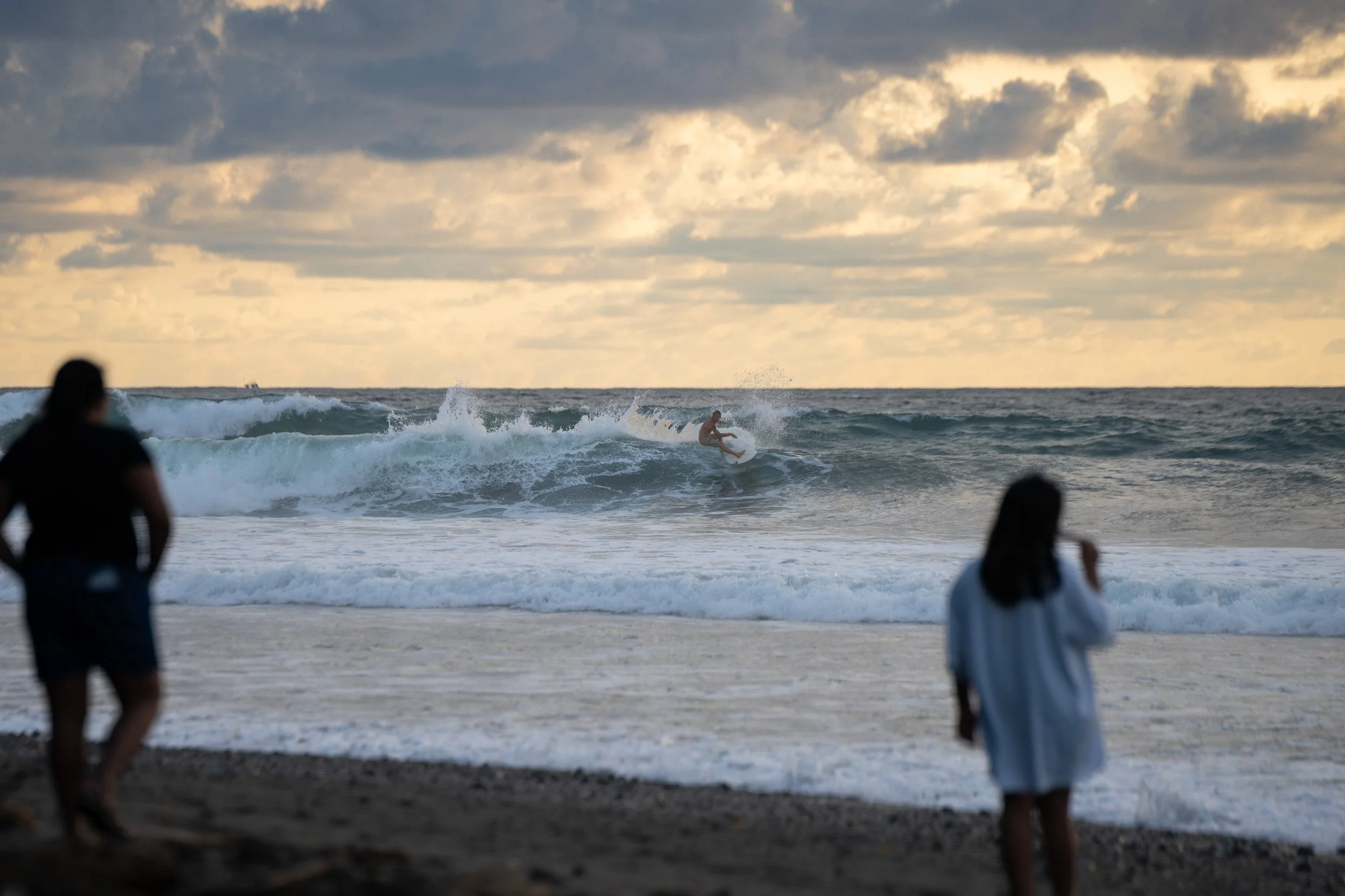 People watching a surfer ride a wave on the beach during cloudy weather, with a distant ship on the horizon.