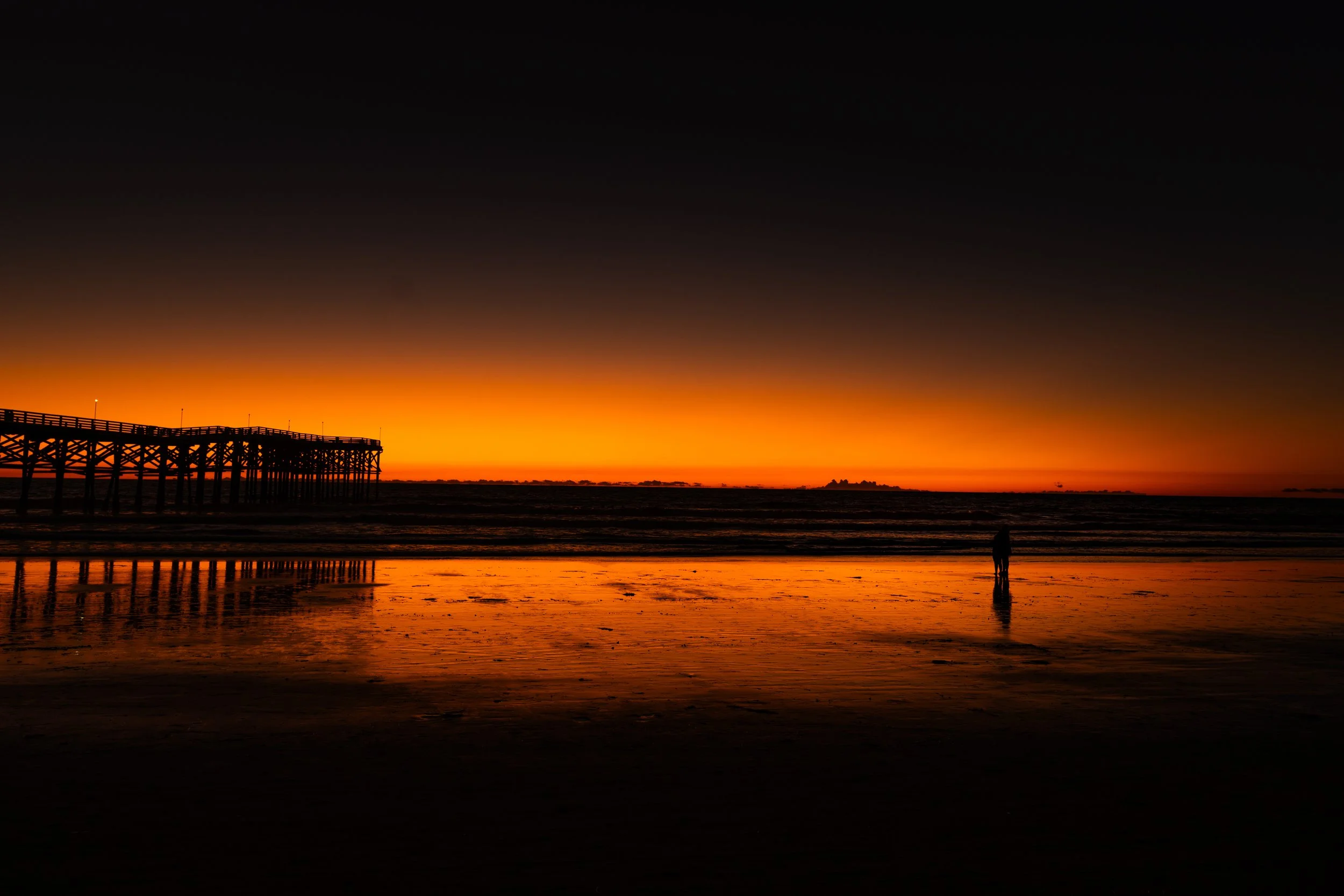 Sunset over the ocean with a pier extending into the water on the left and a person walking along the shoreline on the right, reflected on wet sand.