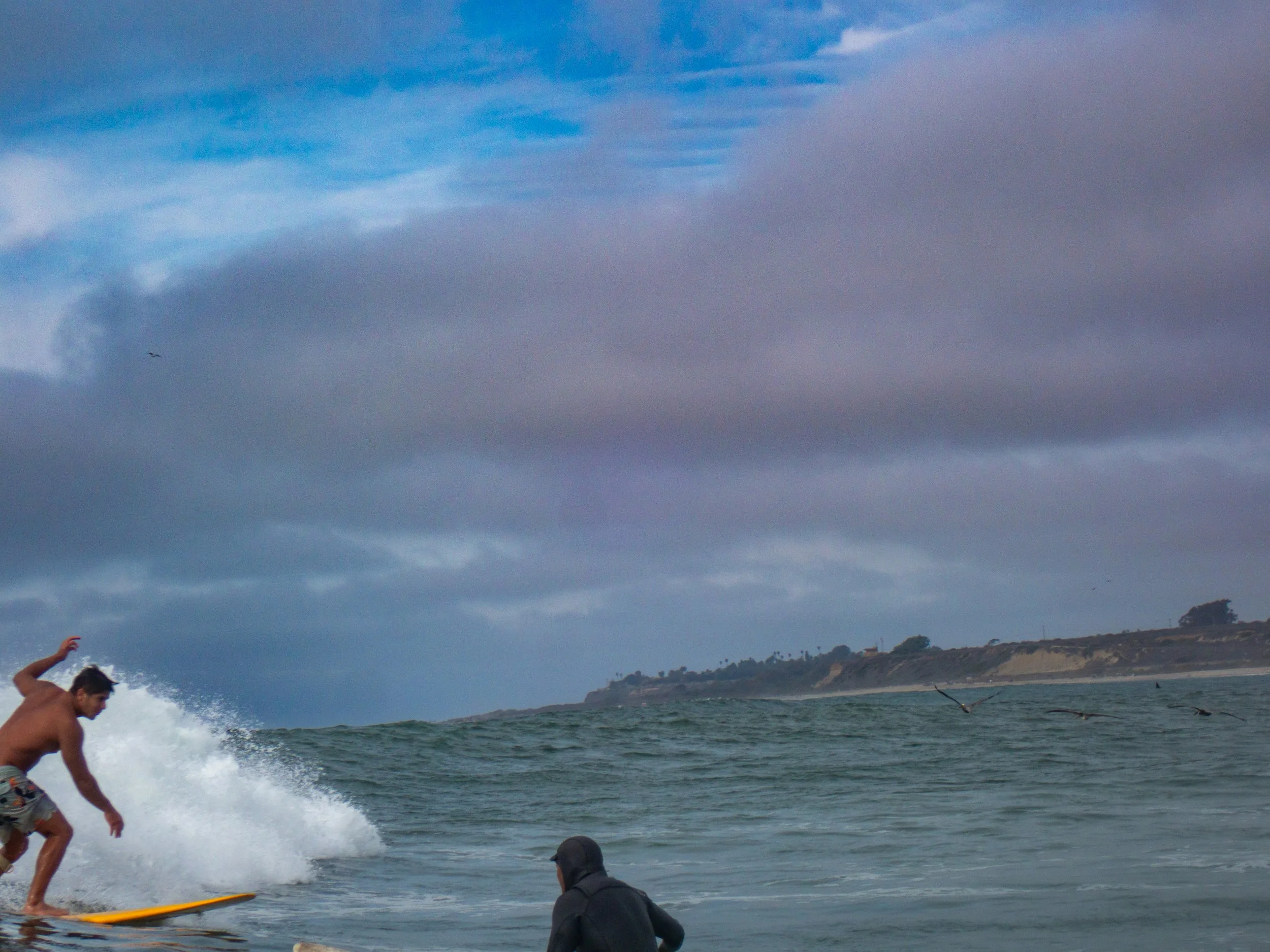 A man surfing on a yellow surfboard in the ocean near a shore with cloudy skies.
