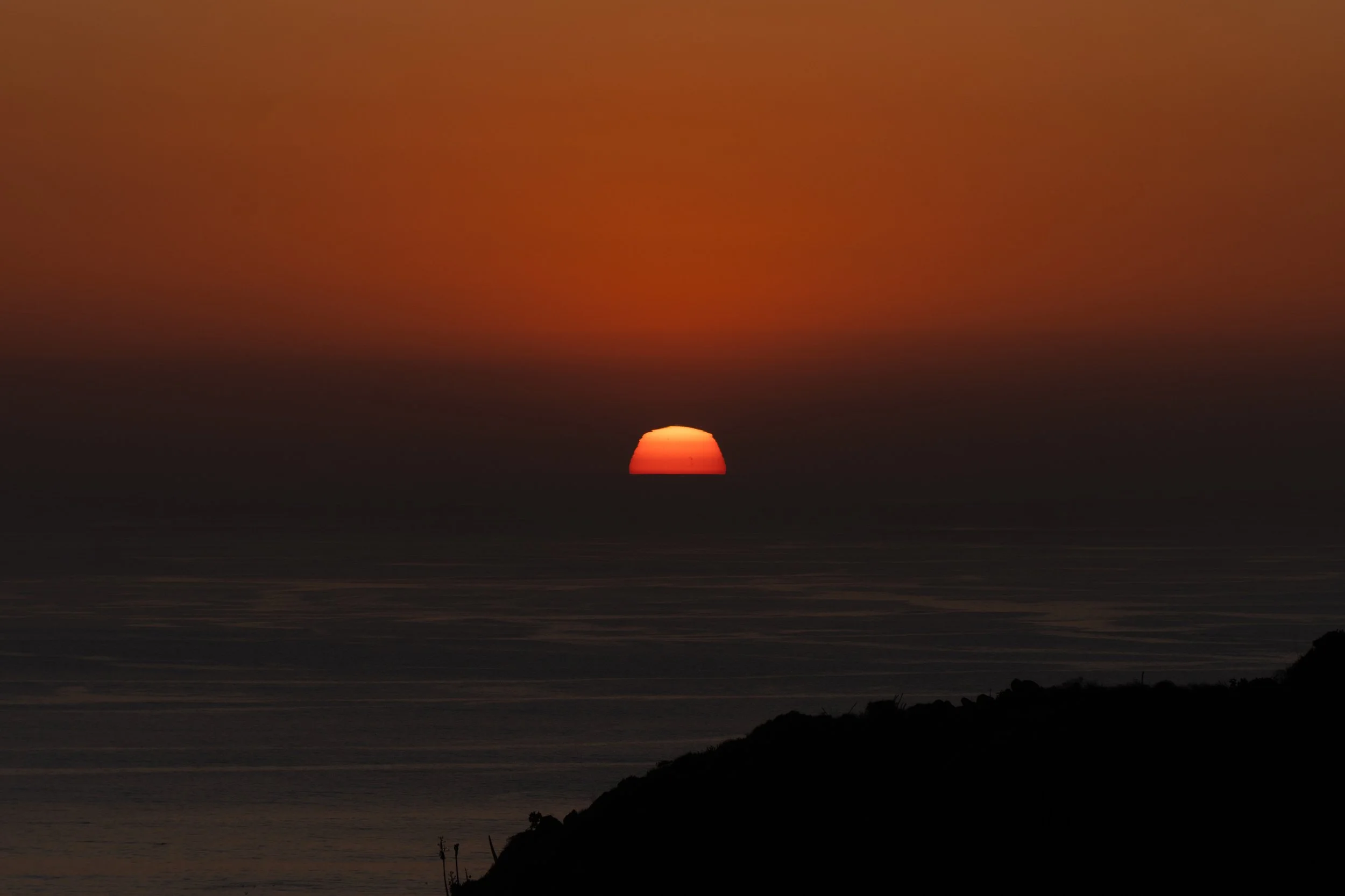 A sunset over the ocean with a reddish-orange sky and a partially hidden sun near the horizon, with a silhouette of land in the foreground.
