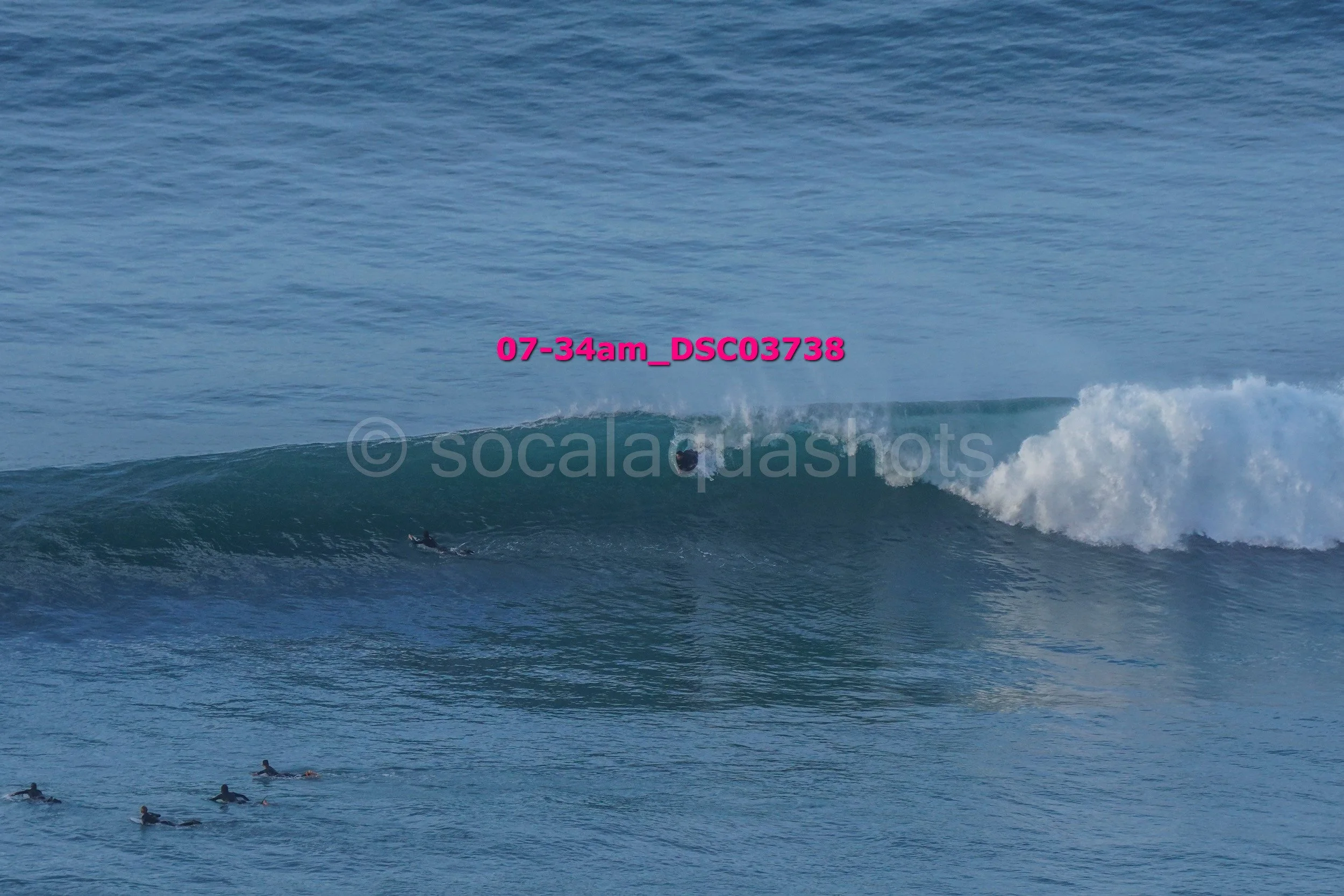 A person surfing a large blue wave in the ocean surrounded by other surfers lying on their surfboards.