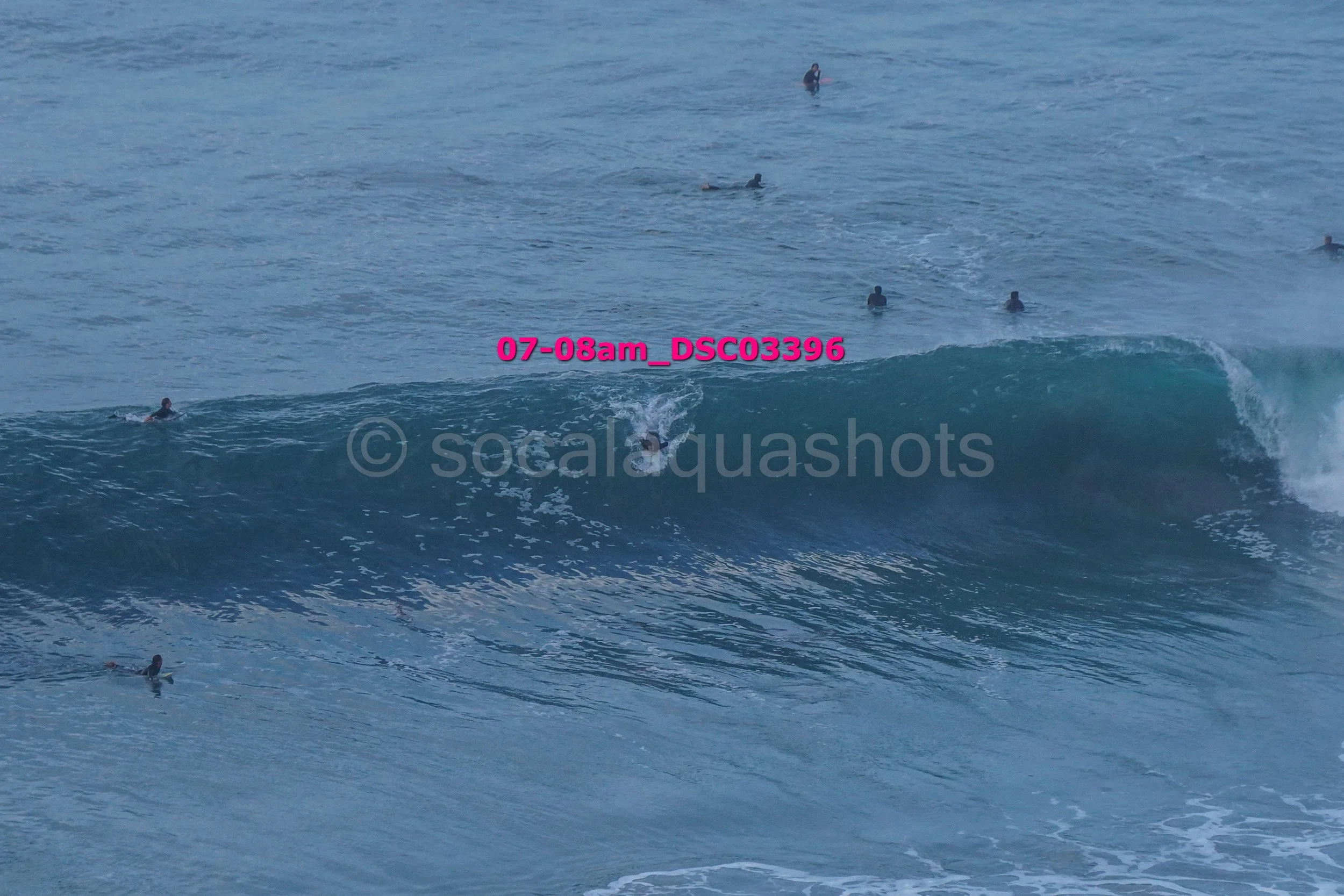 A group of surfers in the ocean, some sitting and some paddling on their boards, with one surfer riding a wave.