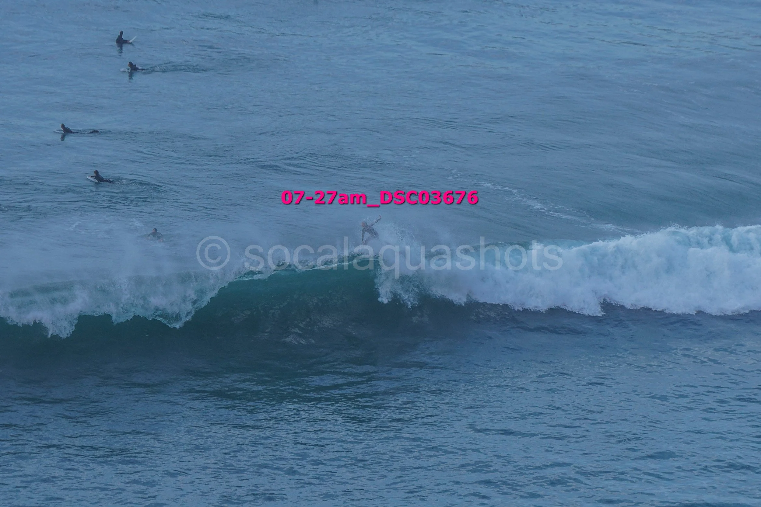 A person surfing on a wave in the ocean, with several other surfers waiting in the water in the background.