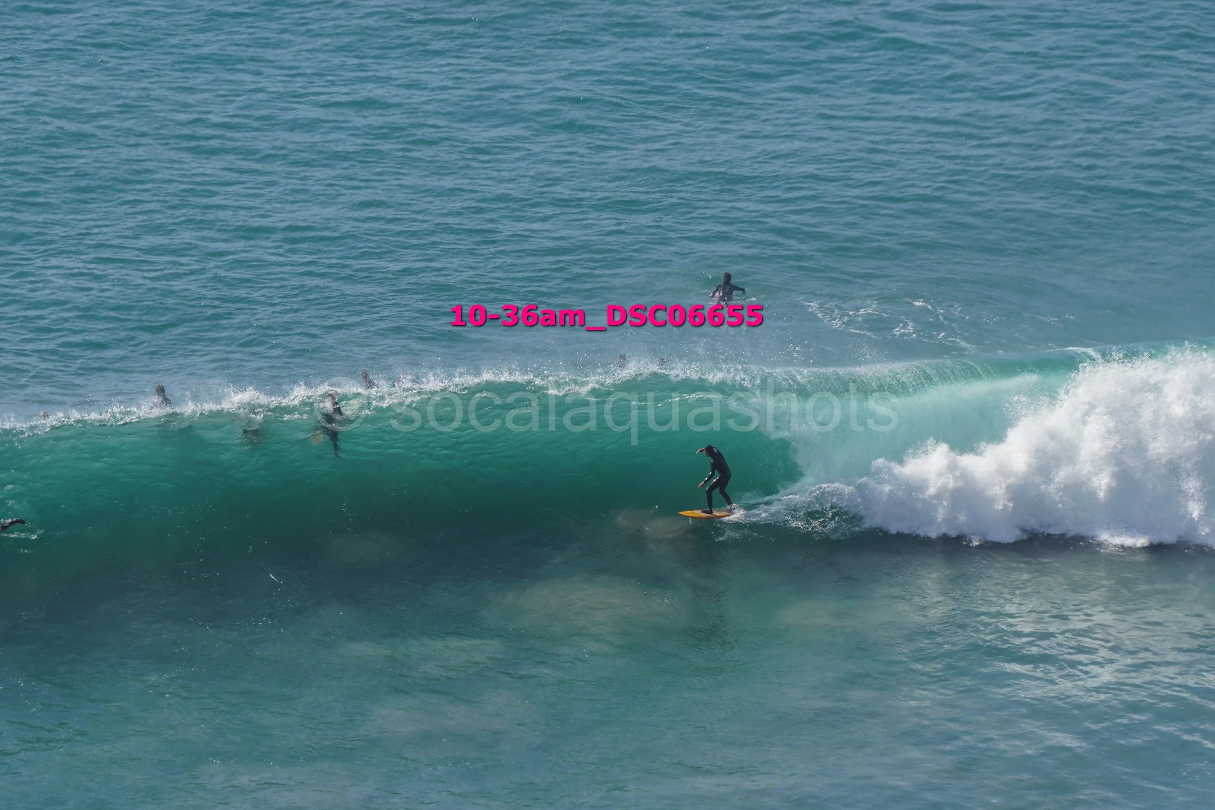 A surfer riding a wave with several other surfers in the water, some swimming and waiting for waves, in an ocean setting.