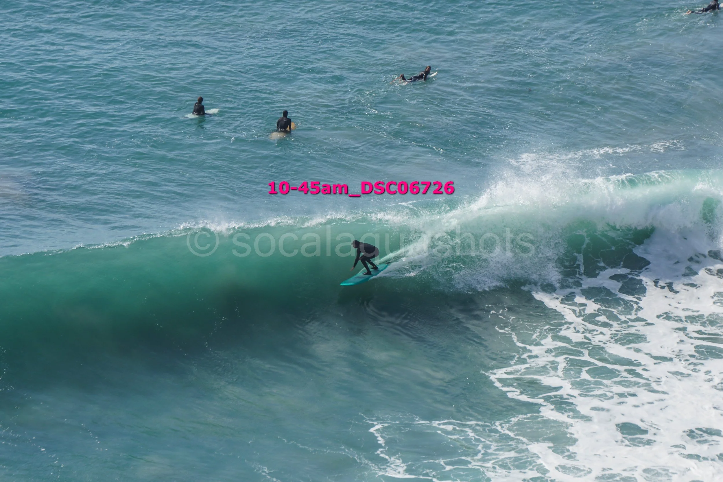 A person surfing on a large ocean wave with five other surfers in the water watching.