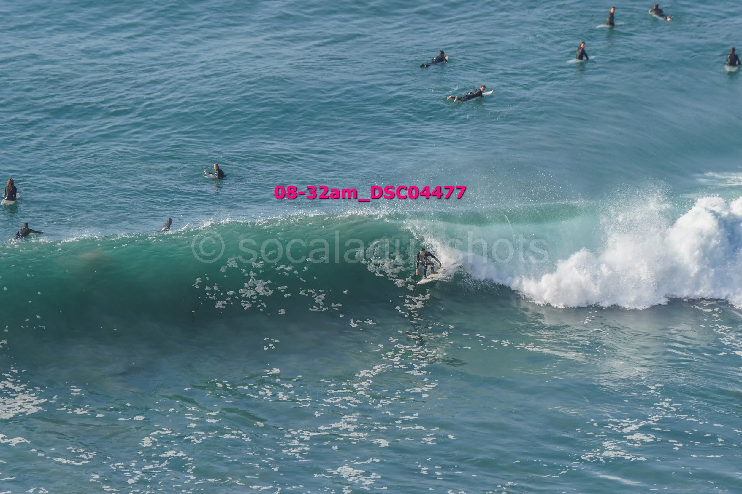Surfer riding a large wave while many surfers wait in the water in the background.
