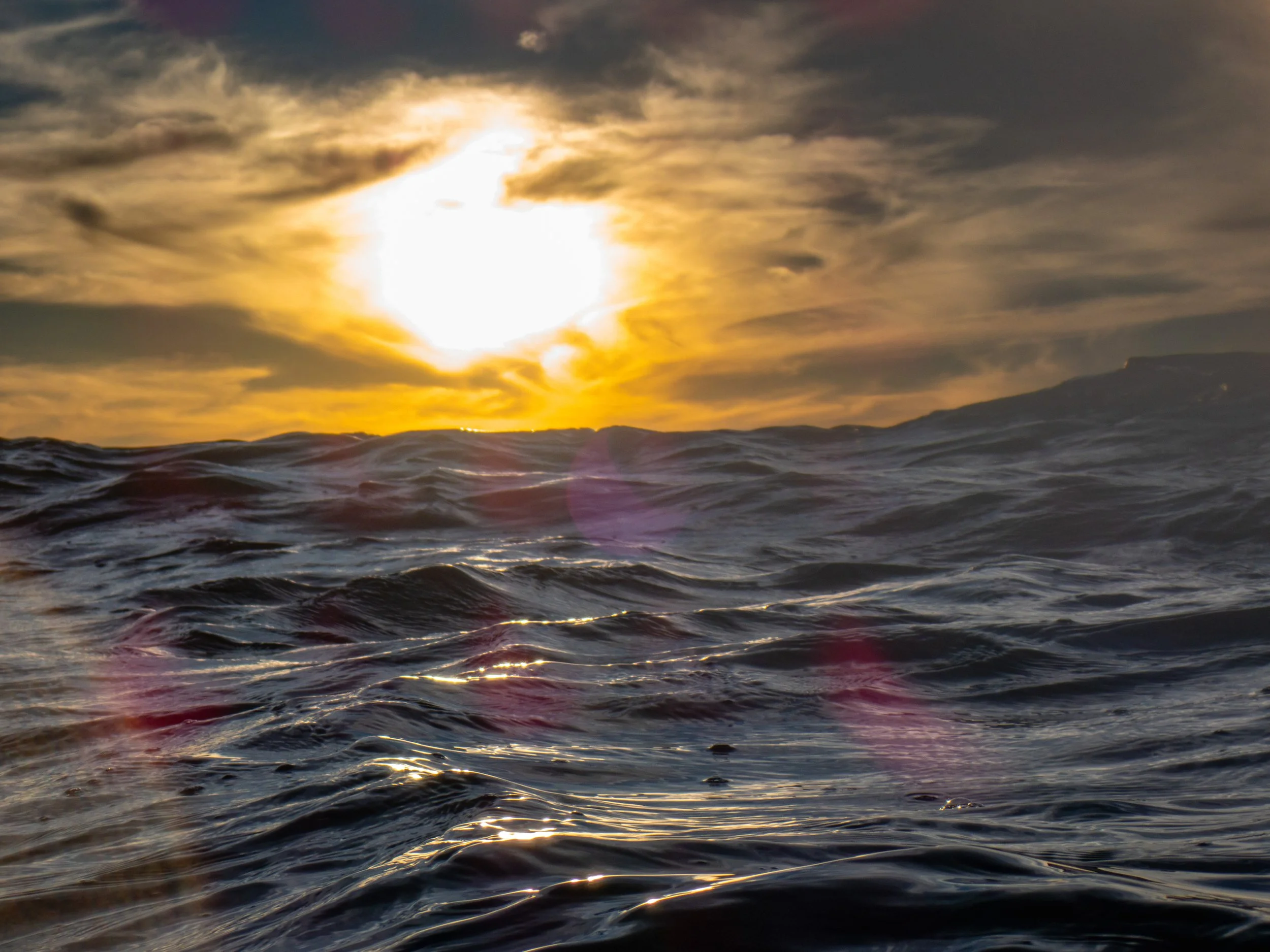 Ocean waves at sunset with the sun partially obscured by clouds in the sky.