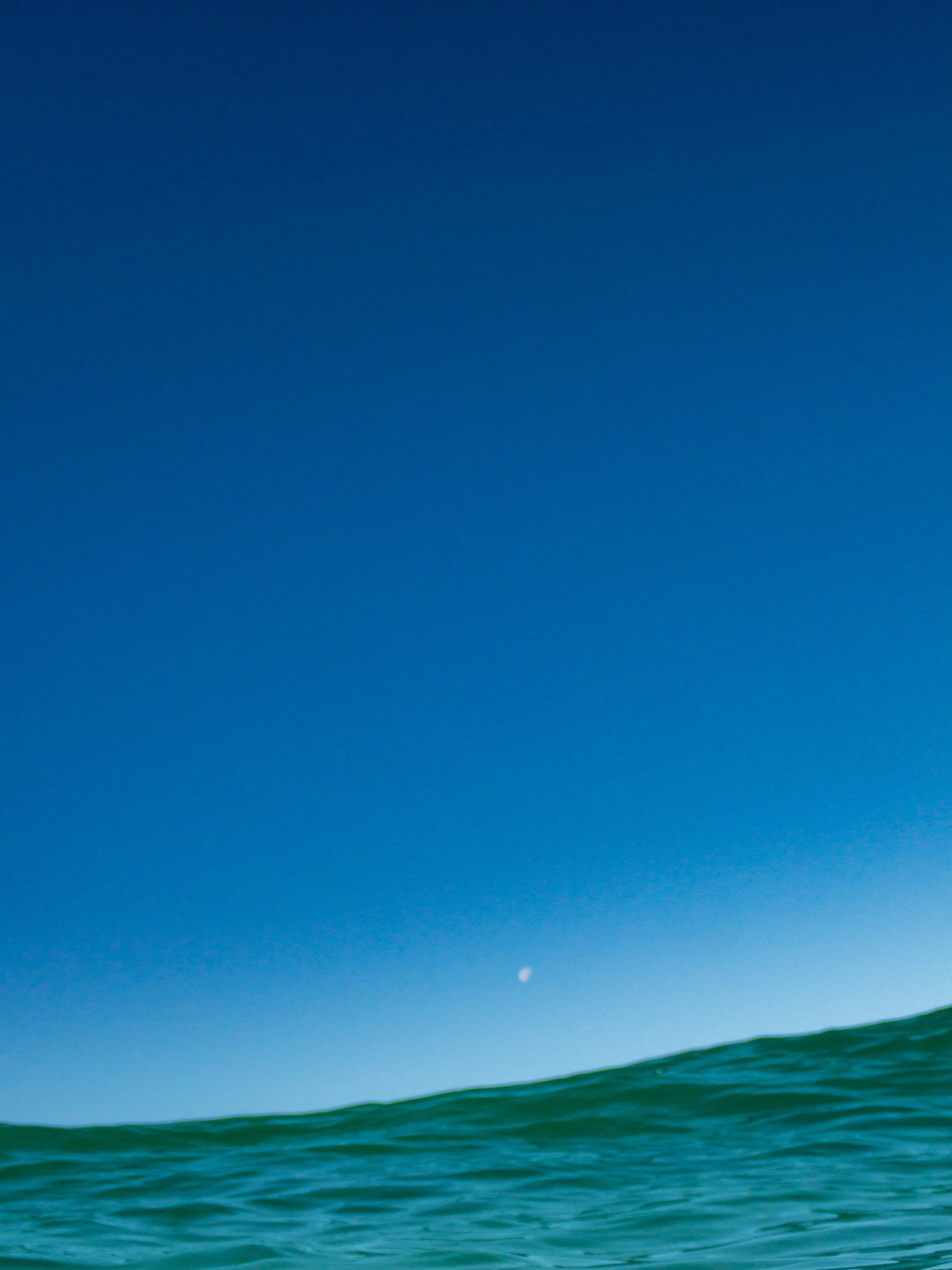Calm ocean waves with a clear blue sky and a small white moon in the distance.