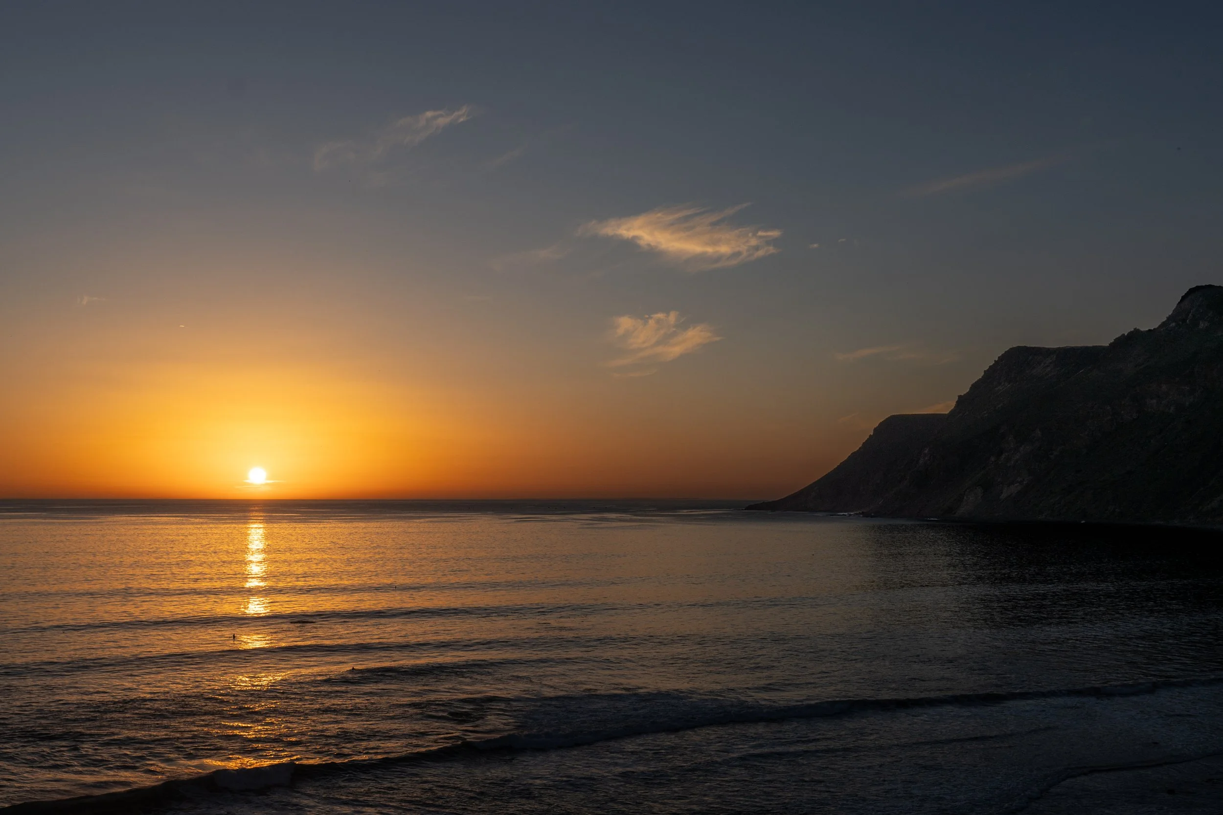 Sunset over the ocean with a mountain on the right and clouds in the sky.