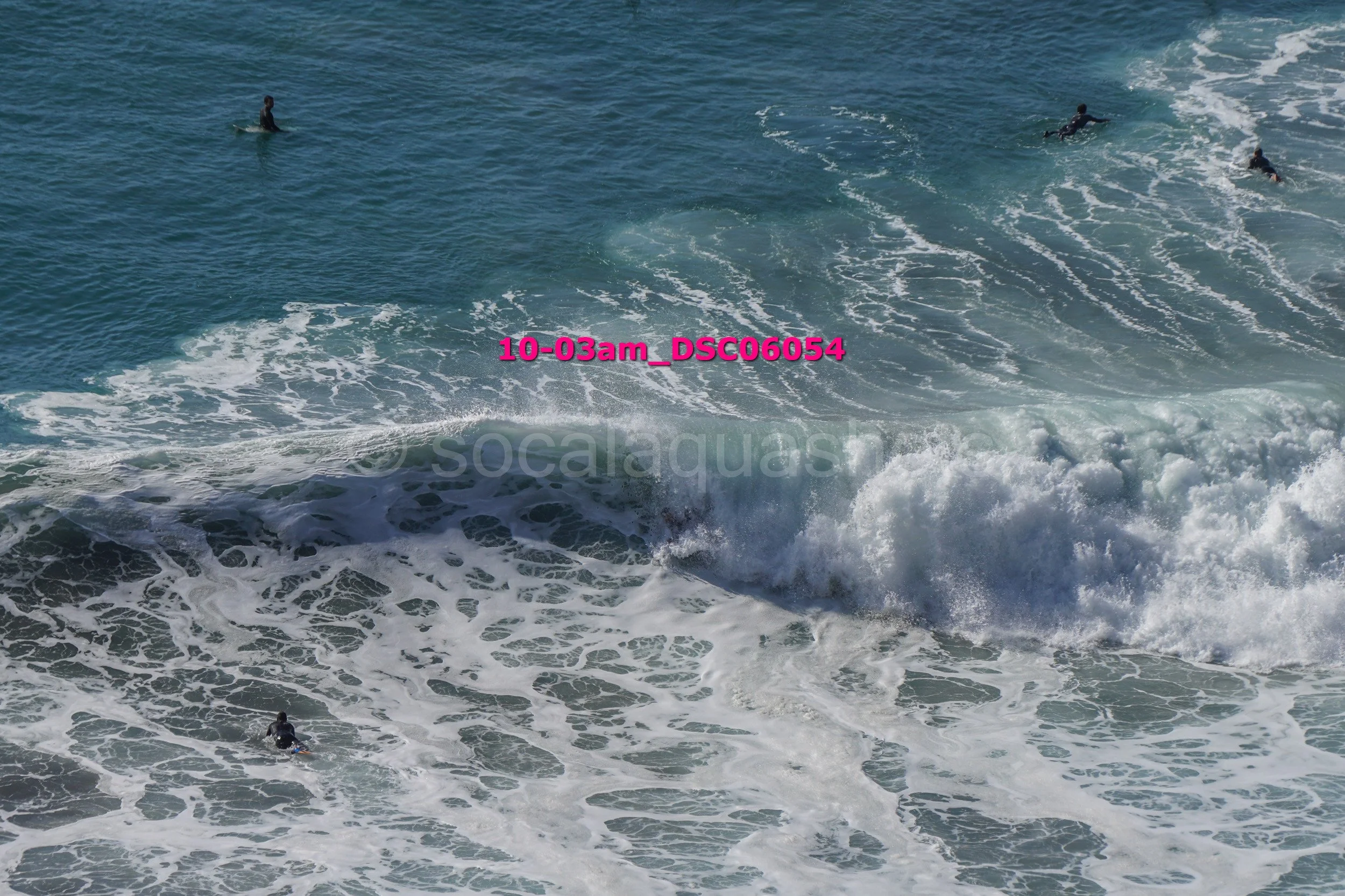Photograph of several surfers riding and waiting for waves in the ocean, with some waves crashing near the shoreline.