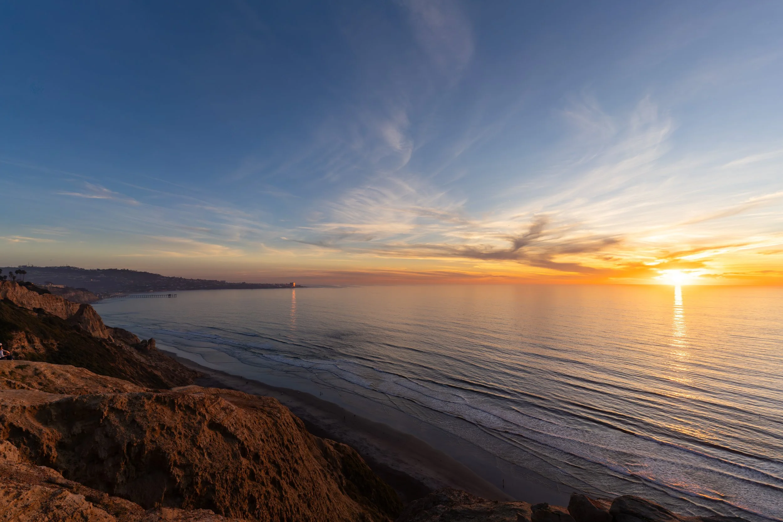 Sunset over the ocean viewed from a rocky cliff, with the sky partly cloudy and the horizon glowing with orange and yellow hues.