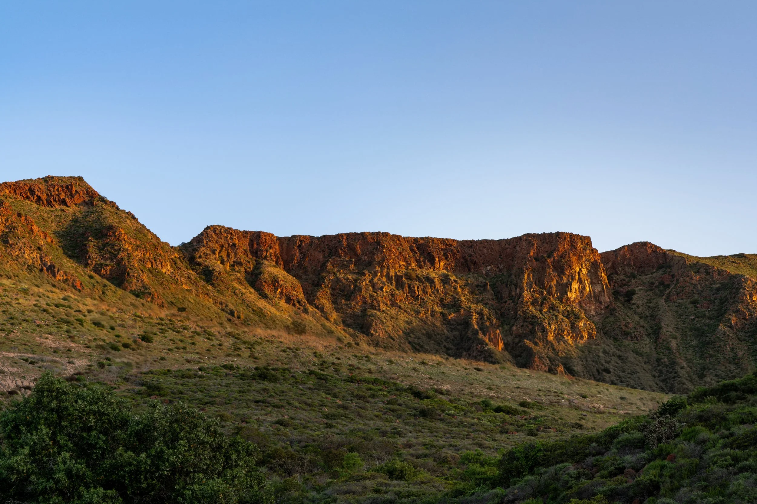 Sunlit mountain range with green vegetation in foreground and clear blue sky in background.