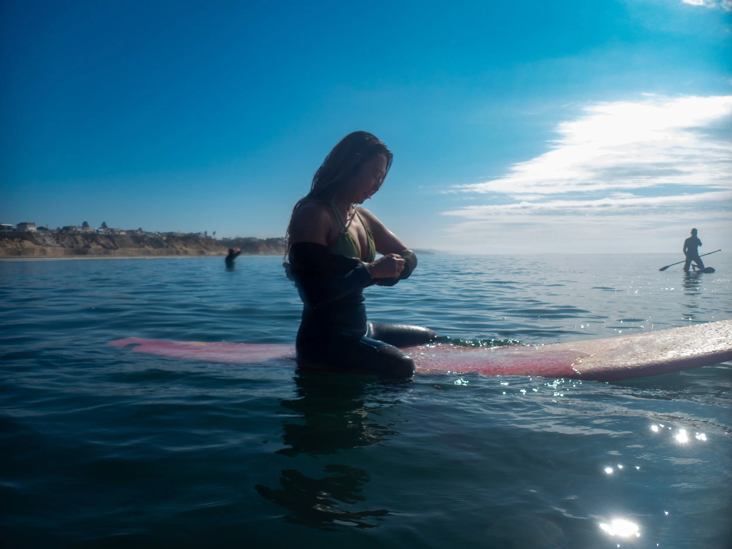 A woman in a black and green swimsuit sitting on her surfboard in the ocean, with a person paddleboarding in the background under a partly cloudy sky.
