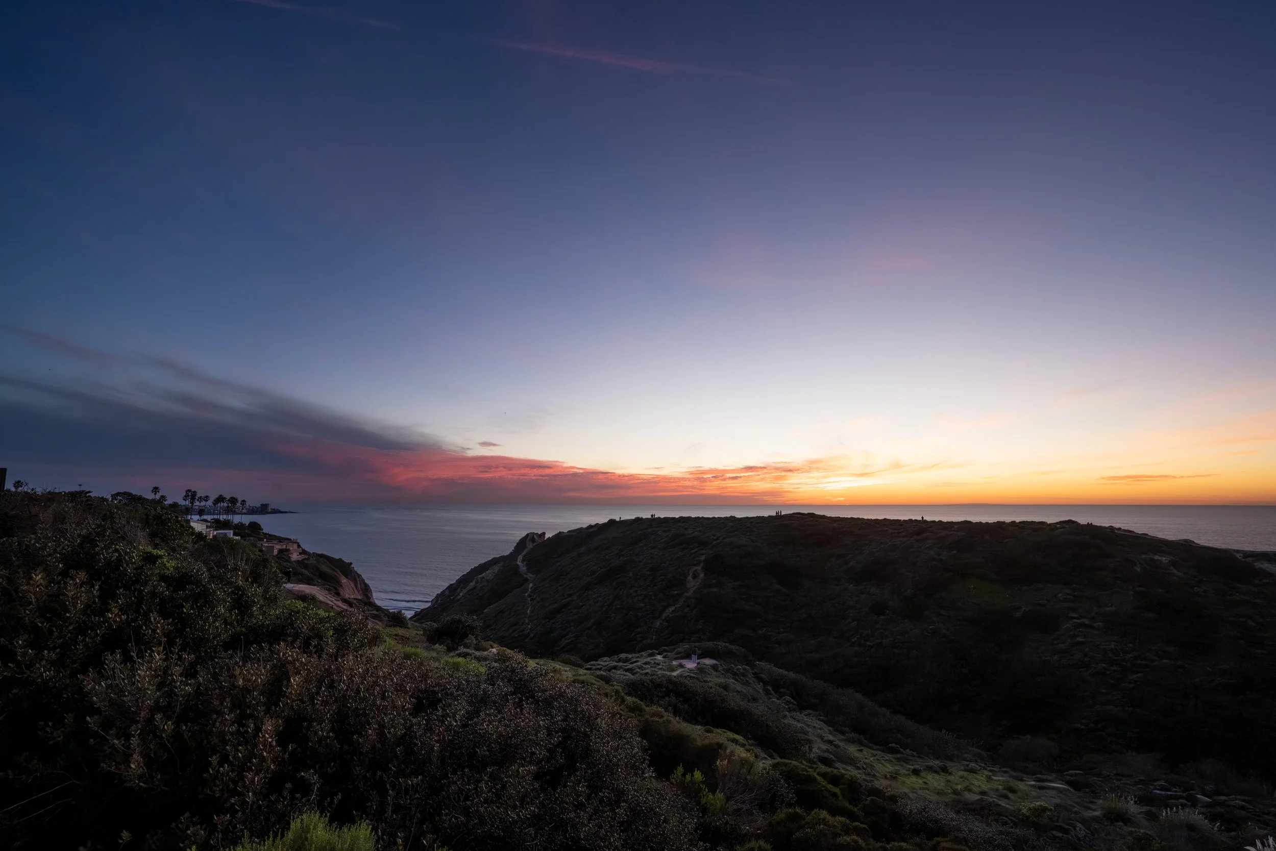 Sunset over a coastal landscape with hills, ocean, and a sky with pink, purple, and orange hues.