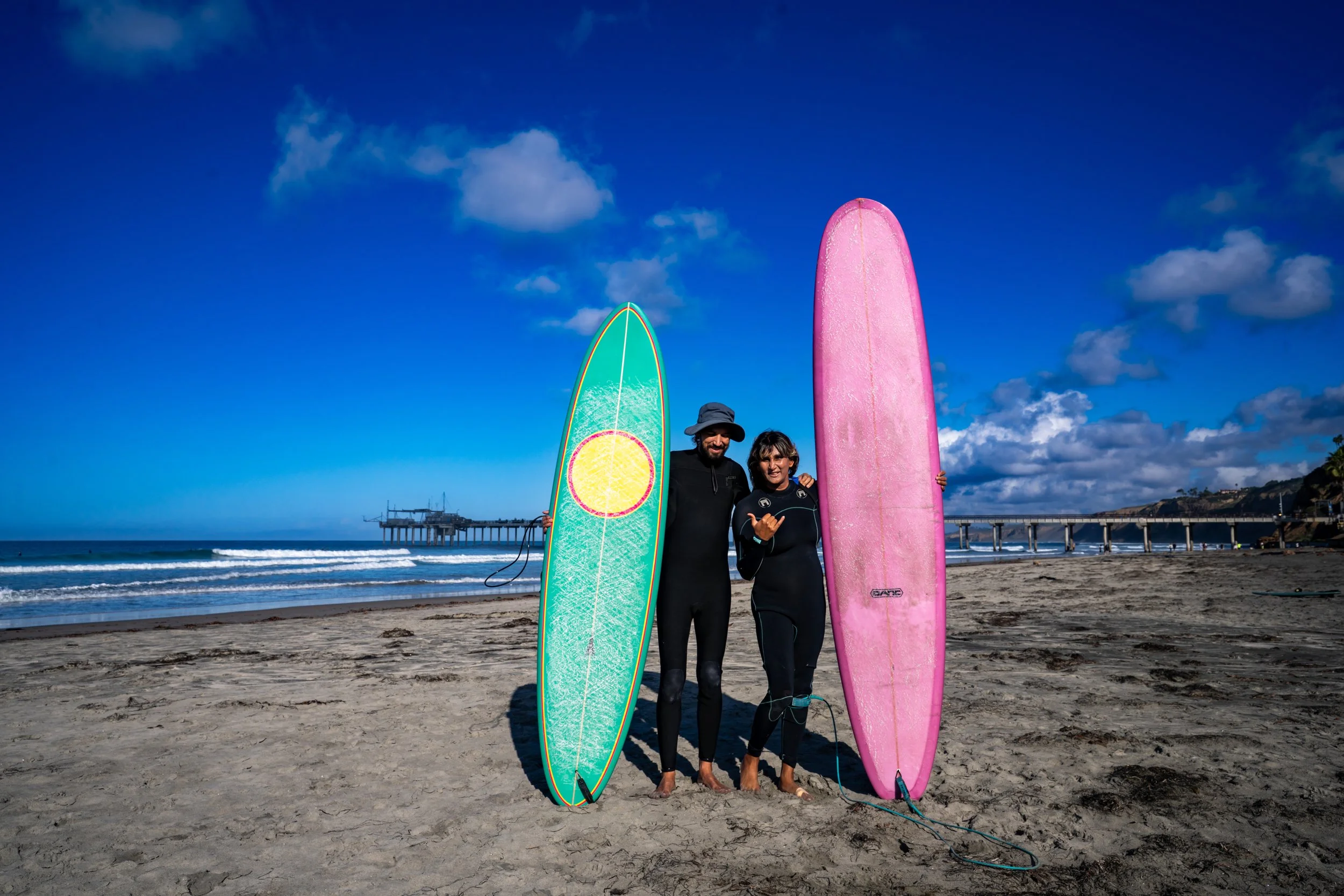 Two people standing on a sandy beach holding surfboards, with a pier and the ocean in the background under a blue sky with scattered clouds.