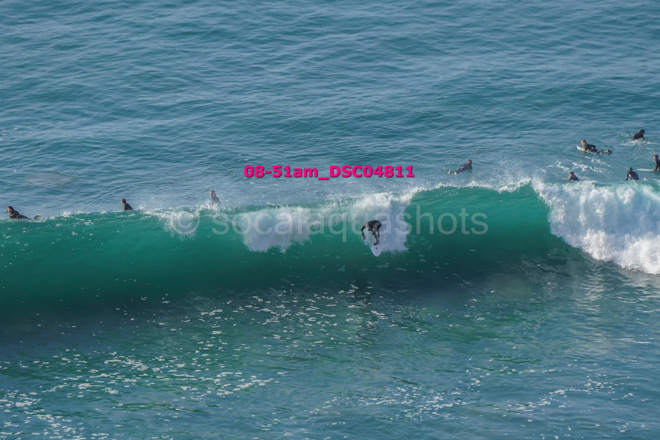 Surfer riding a wave while multiple people in wetsuits watch from the water in the background.