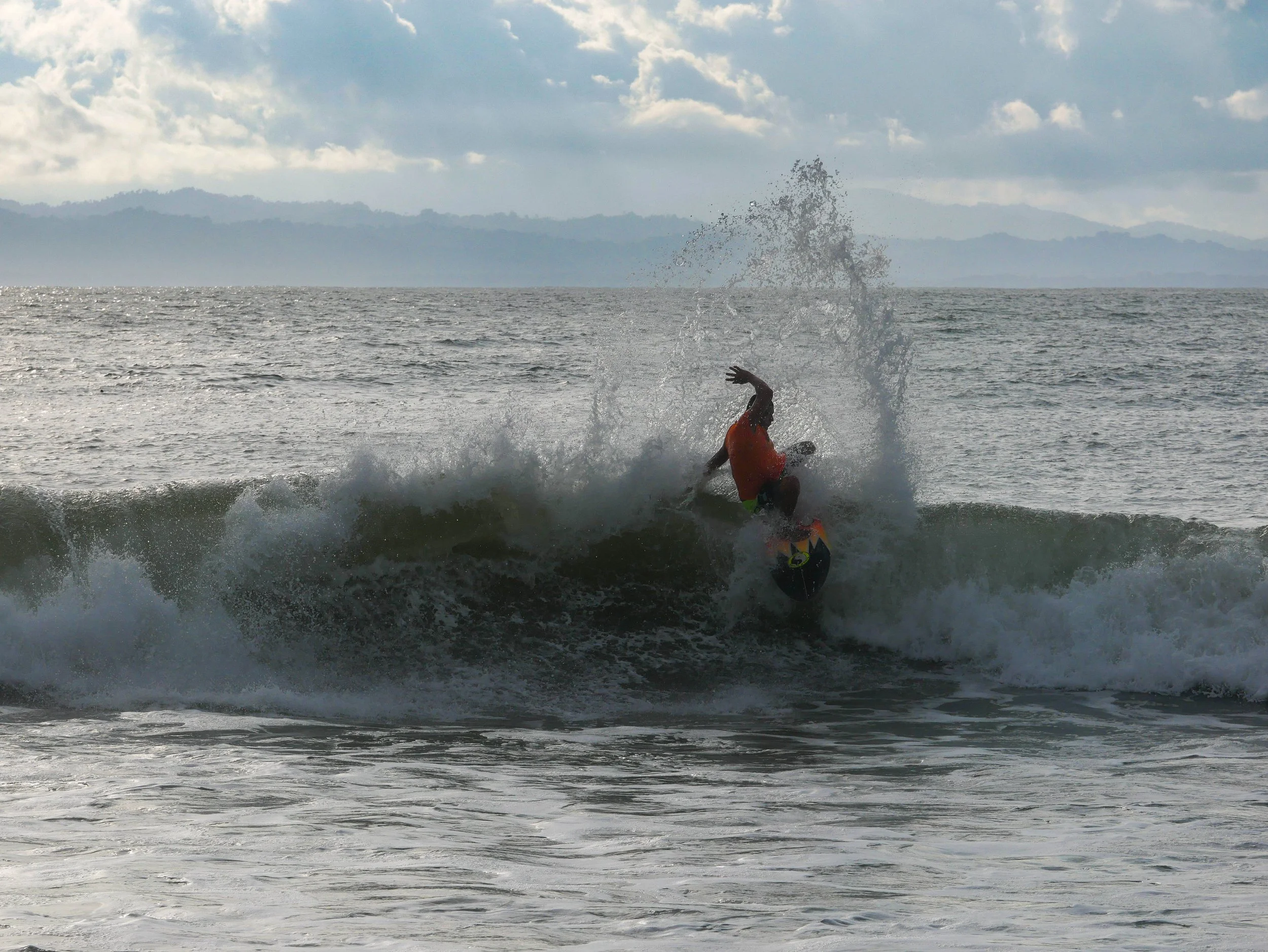 Surfer riding a wave in the ocean with a cloudy sky and distant mountains in the background.
