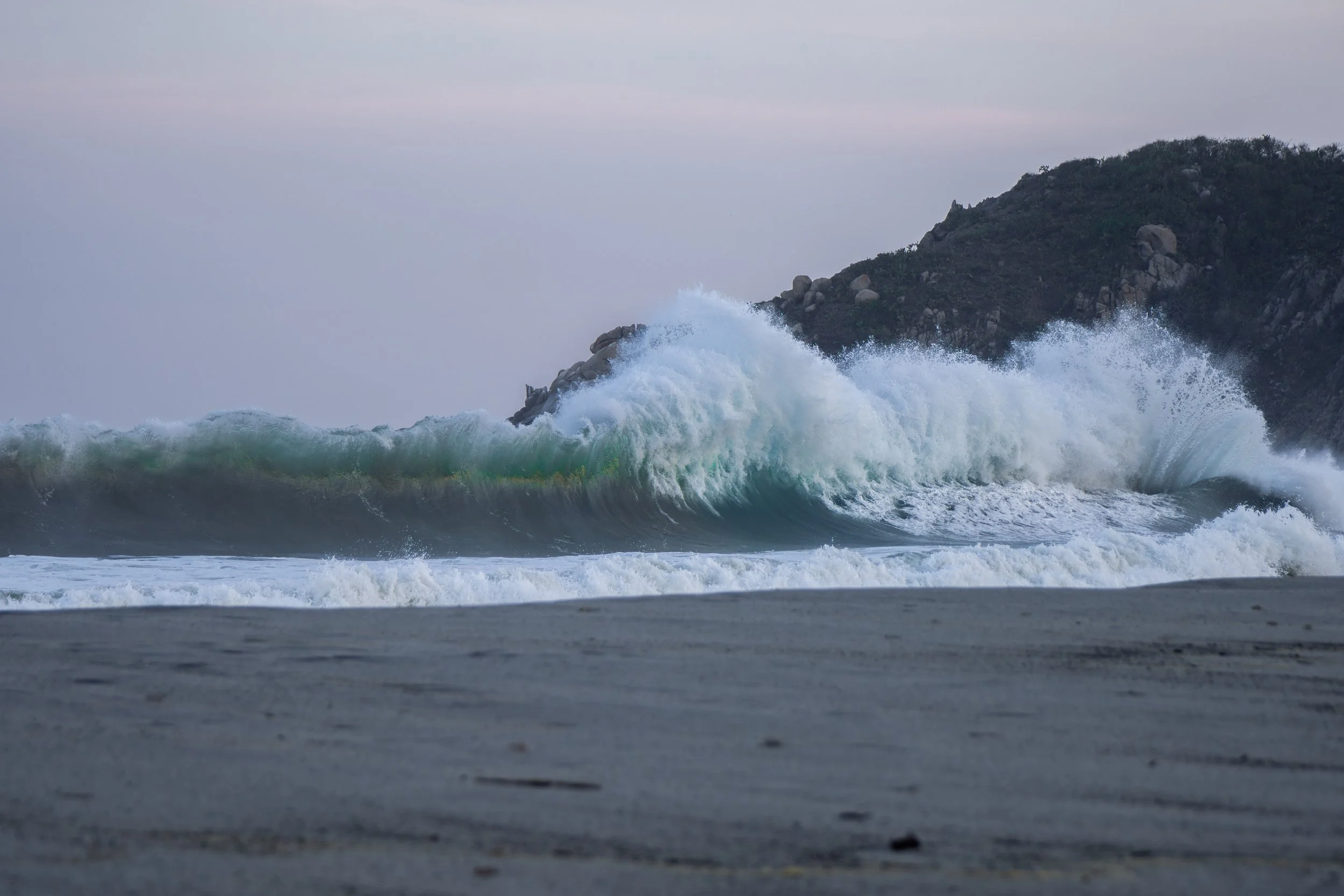Ocean waves crashing against rocks near a hillside with sparse vegetation under a cloudy sky.